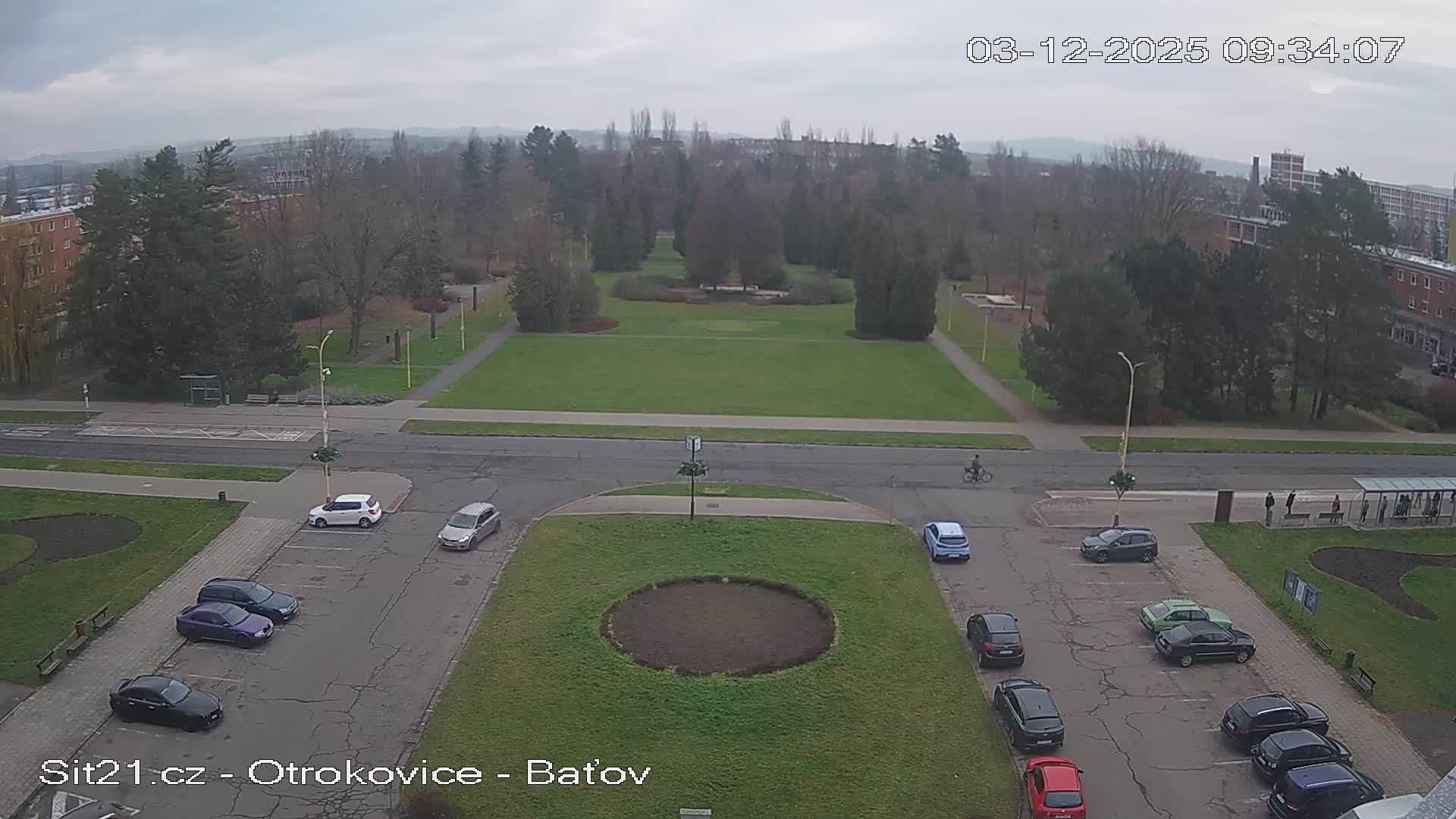 An elevated view showcases an urban landscape featuring a large green park with paths and numerous trees, flanked by roads with parked cars and multi-story buildings, all beneath a uniform, overcast sky.