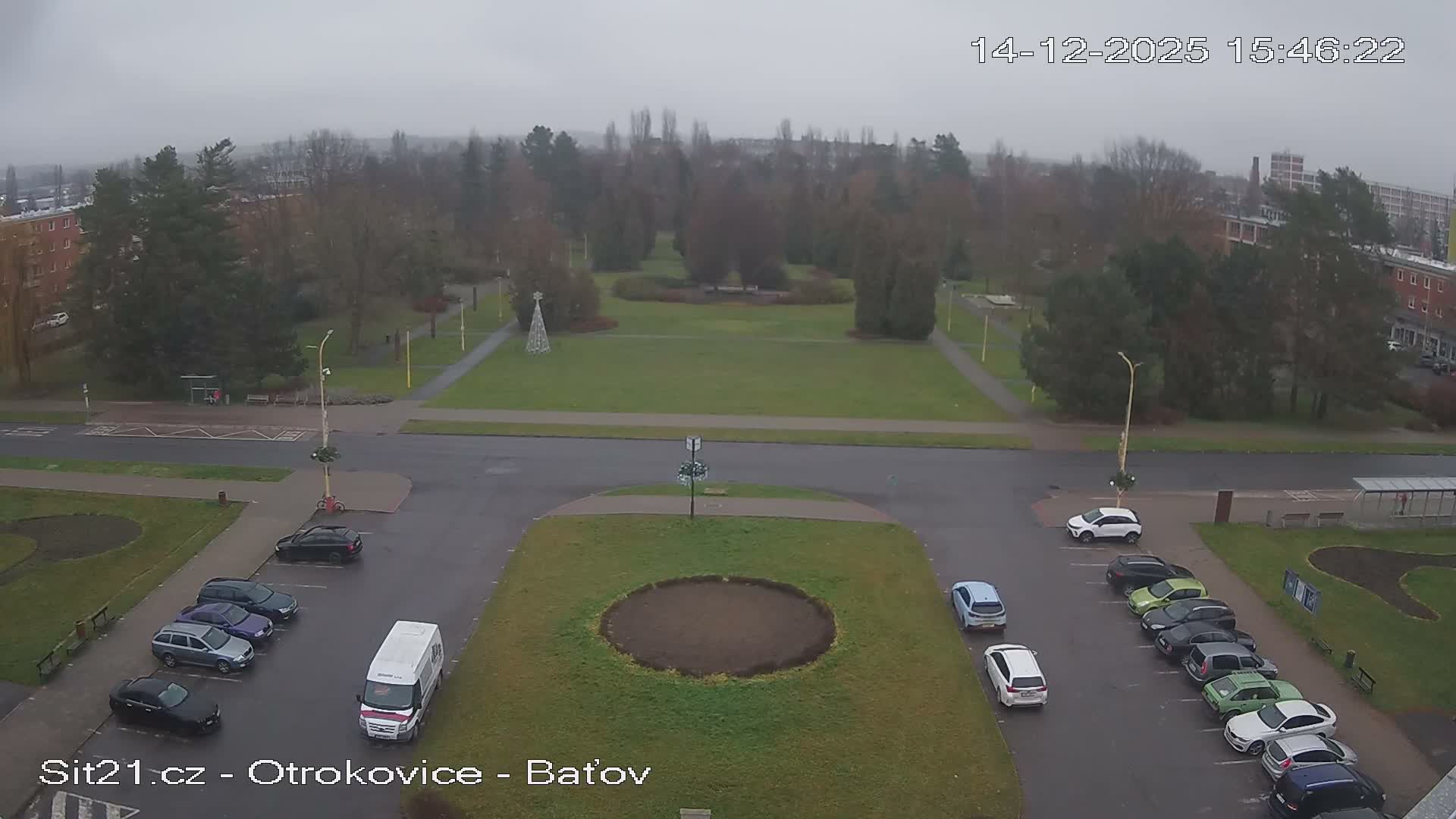 An elevated view showcases an urban landscape featuring a large green park with paths and numerous trees, flanked by roads with parked cars and multi-story buildings, all beneath a uniform, overcast sky.