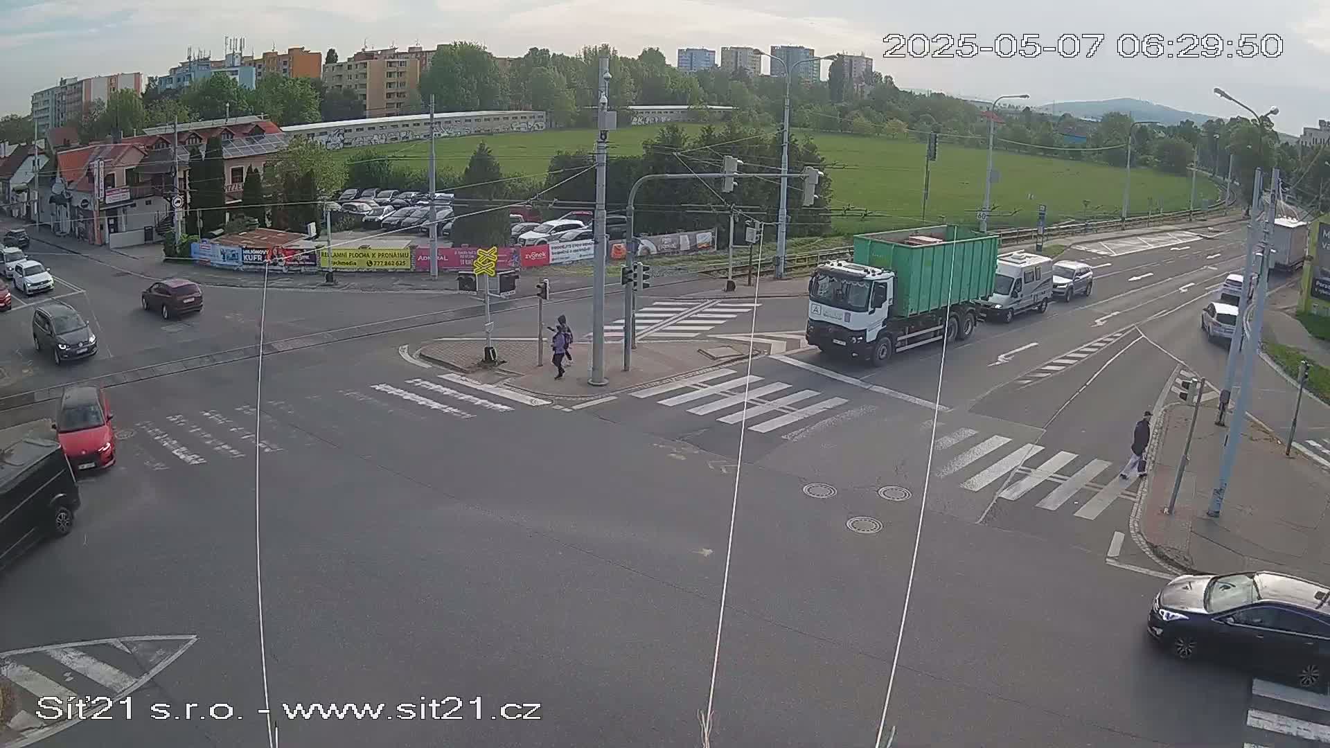 A partly cloudy day reveals a busy intersection with several cars, a garbage truck, and pedestrians crossing crosswalks.
