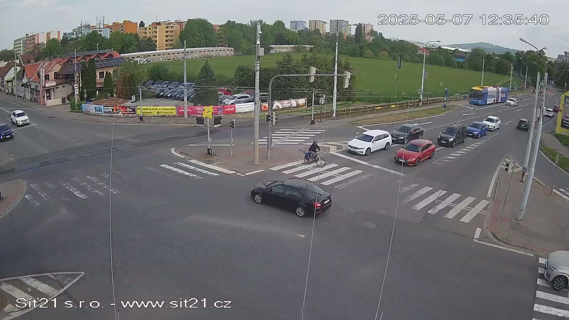 Several cars and a bus are driving through a multi-lane intersection on a partly cloudy day, with a cyclist crossing a crosswalk.