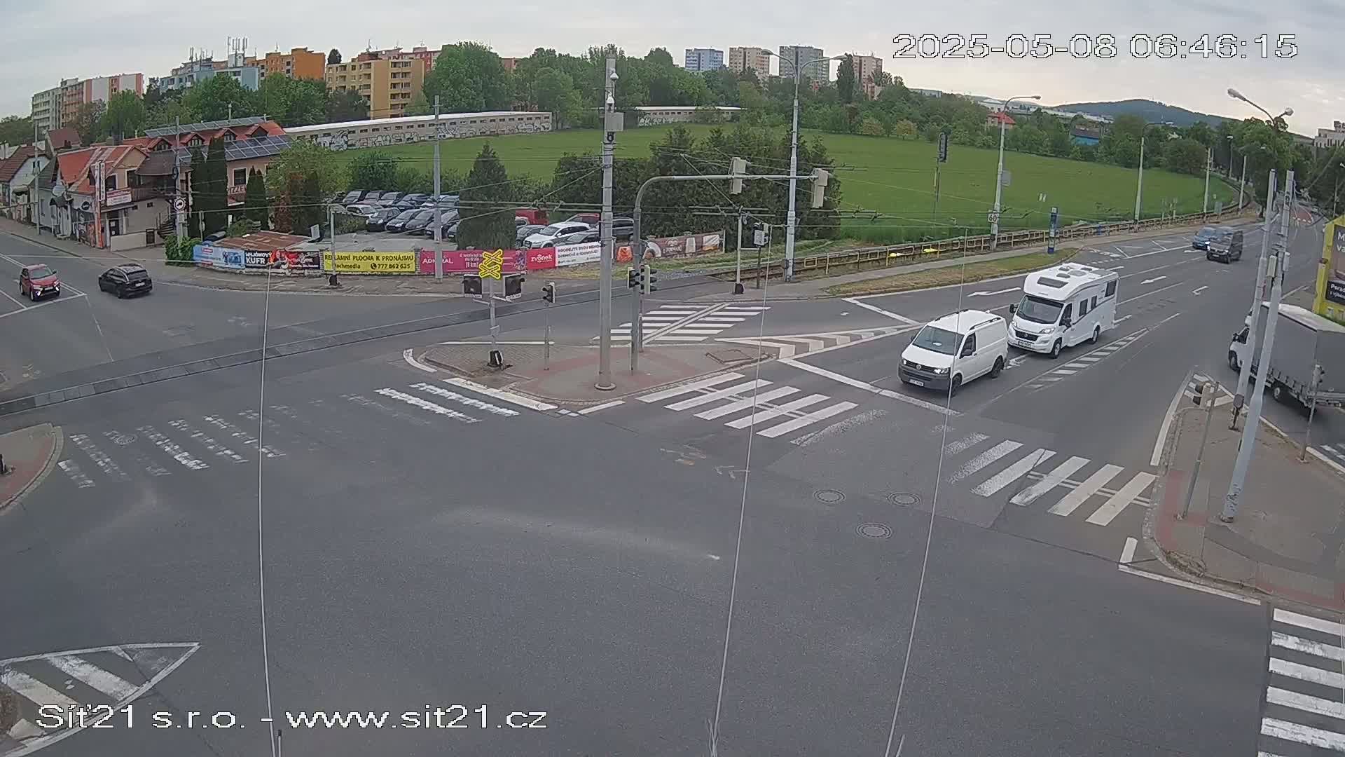 A mostly sunny overhead view of a street intersection with several vehicles, including vans and a campervan, navigating crosswalks and traffic signals.