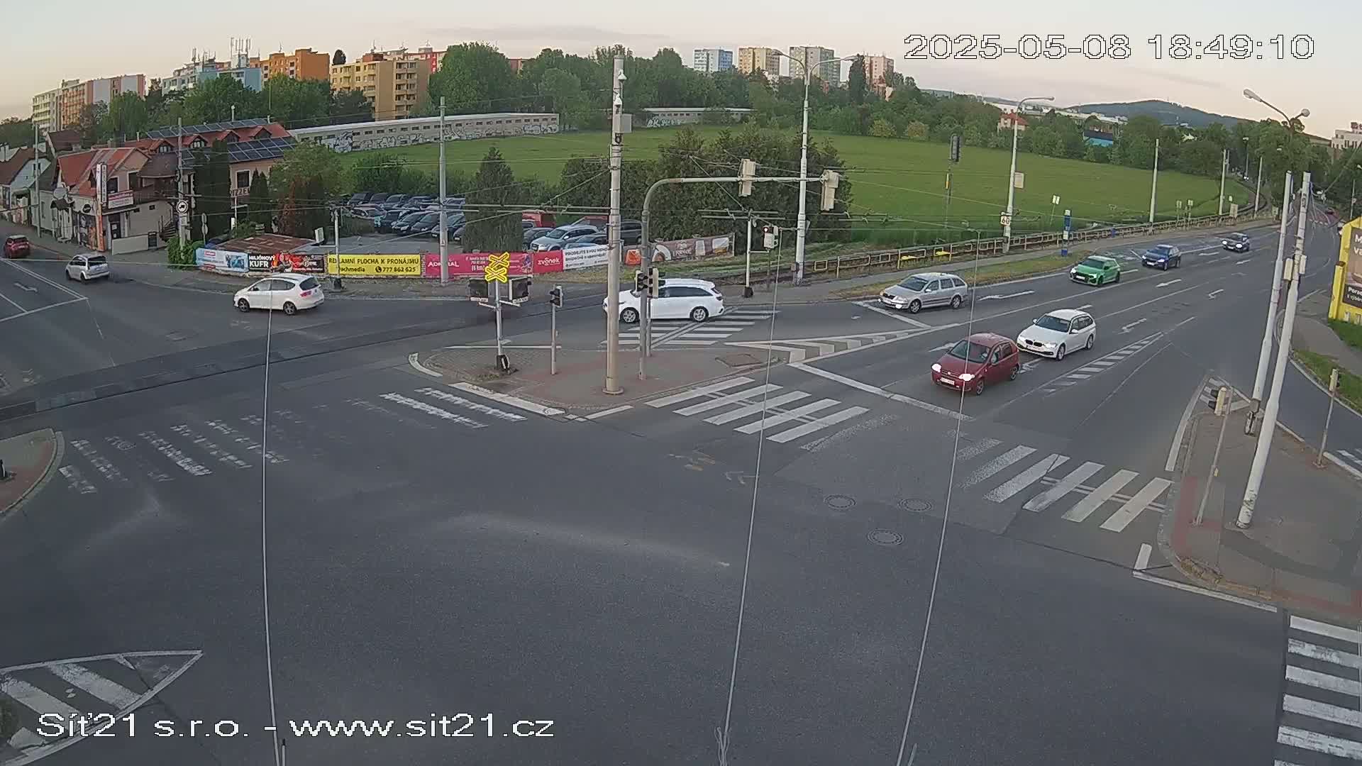Several cars are driving through a multi-lane intersection at dusk, with a grassy field and buildings visible in the background.