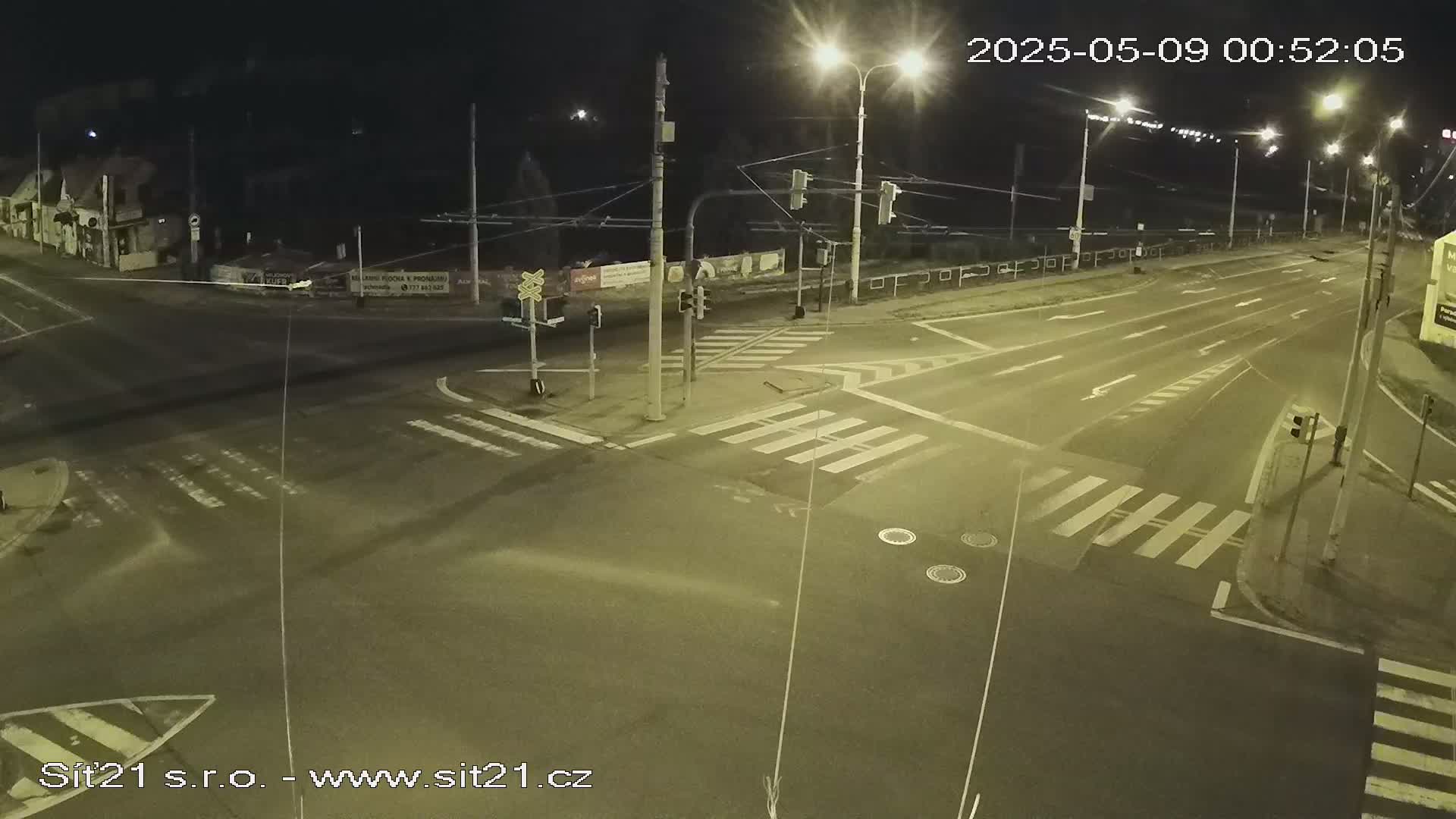 An empty, multi-lane road intersection at night, illuminated by streetlights, with pedestrian crosswalks and traffic signals.
