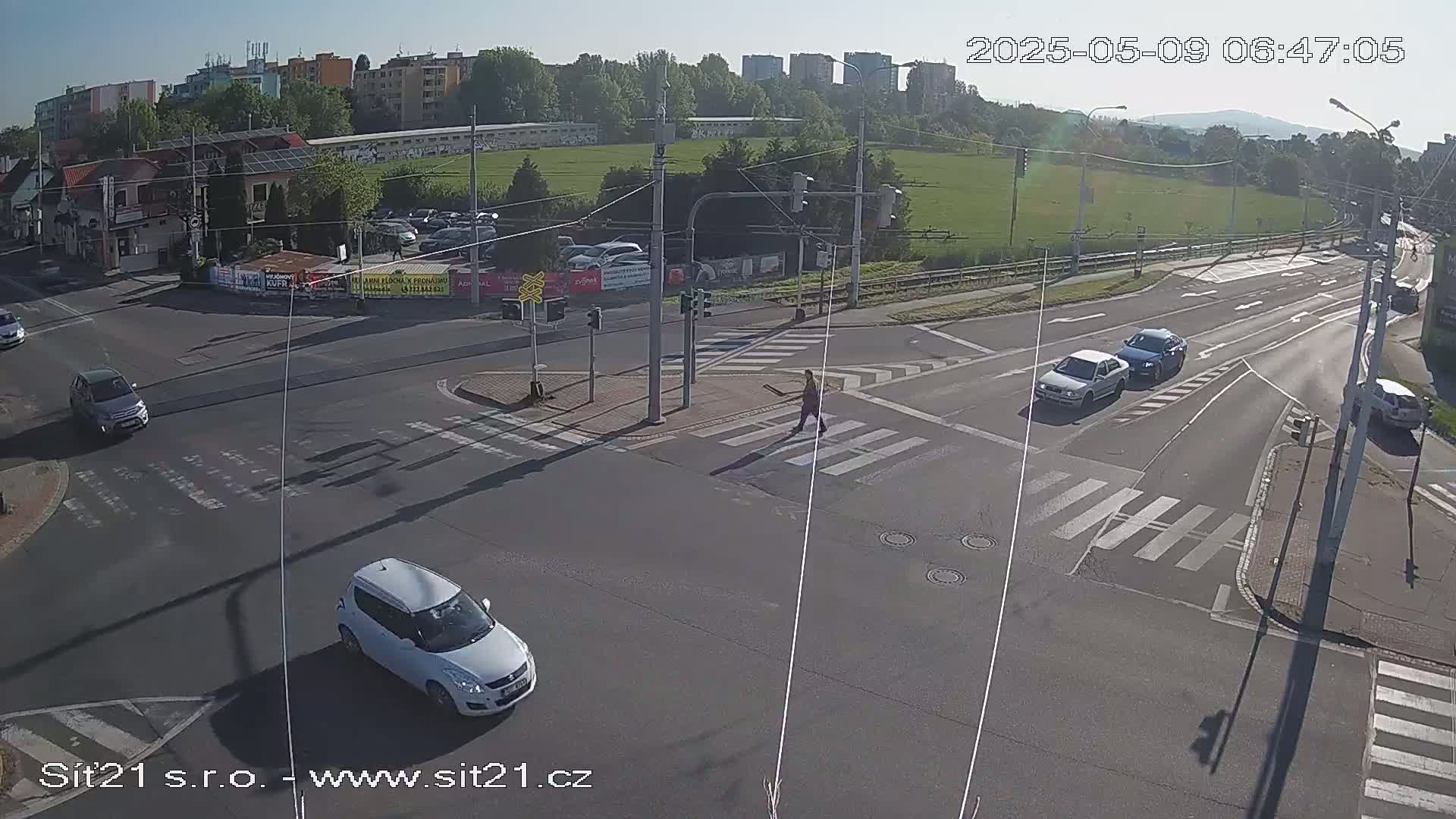 A sunny, high-angle view of a multi-lane road intersection with several cars, a pedestrian crossing, and buildings in the background.