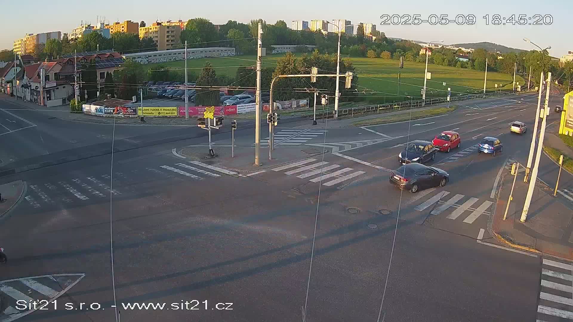 Several cars are navigating a multi-lane intersection with crosswalks under a partly sunny evening sky.