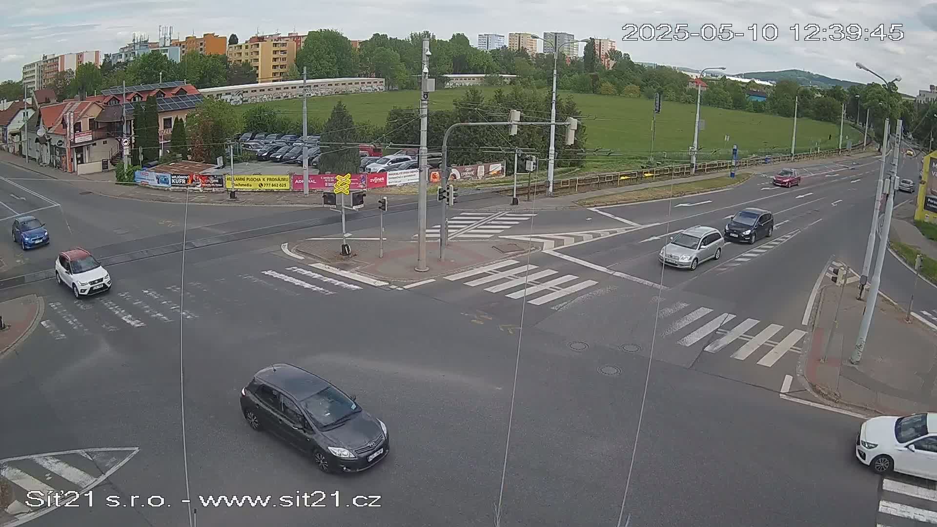 Several cars are navigating a multi-lane intersection with pedestrian crosswalks under a partly cloudy sky.