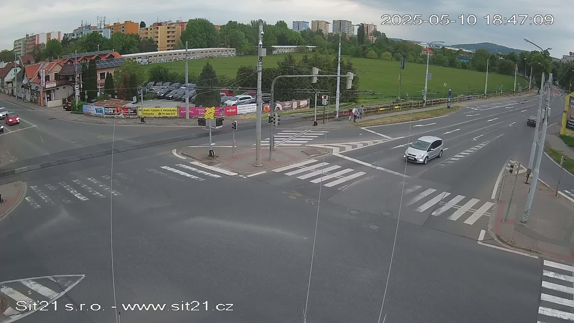 A mostly empty street intersection with crosswalks and a silver minivan driving through under partly cloudy skies.