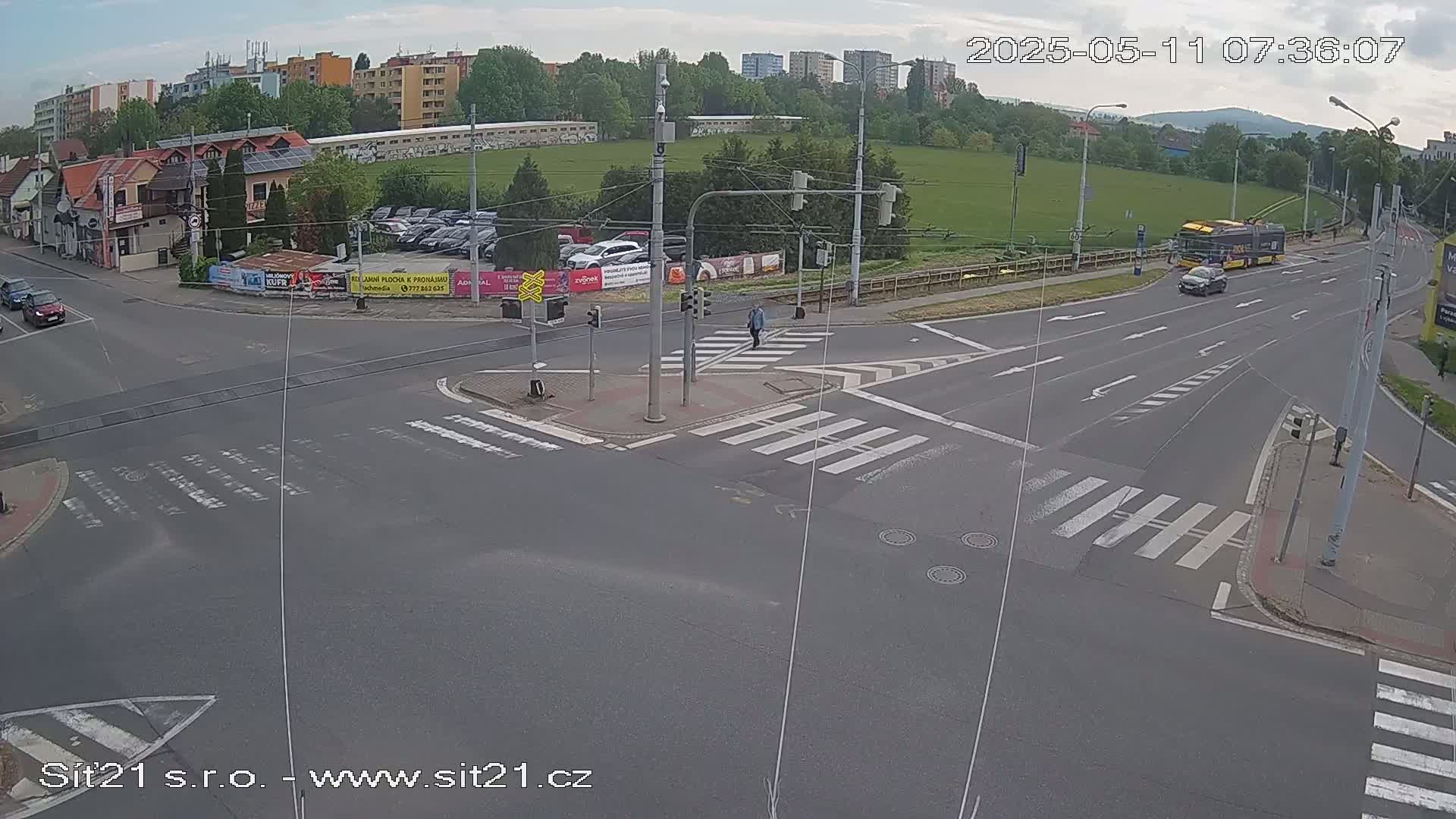 A mostly sunny aerial view of a multi-lane road intersection with crosswalks, pedestrians, and several vehicles including a bus.