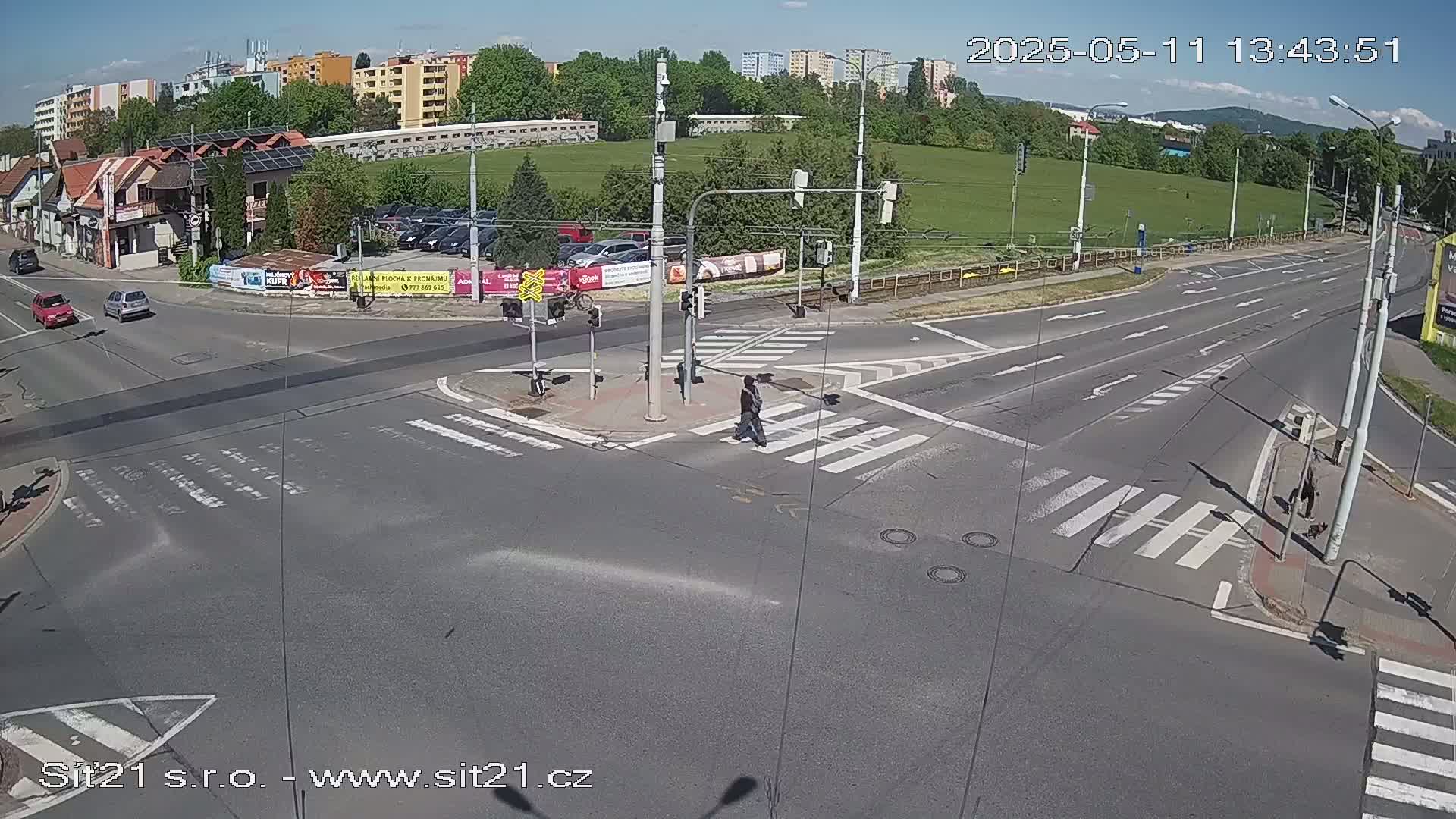 A sunny day at a multi-lane road intersection with pedestrians crossing, several cars at the intersection, and buildings and a grassy field in the background.