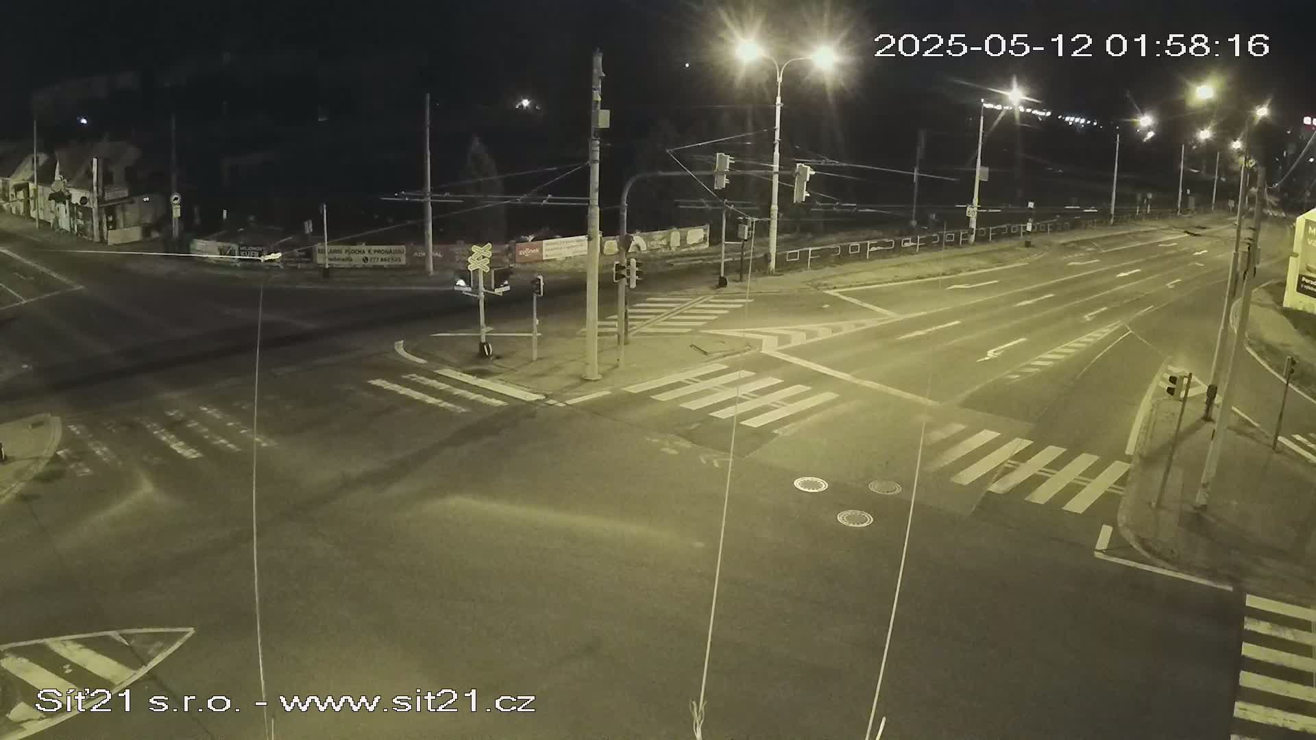 A nighttime view of a multi-lane road intersection with crosswalks, traffic signals, and overhead wires, under clear skies.