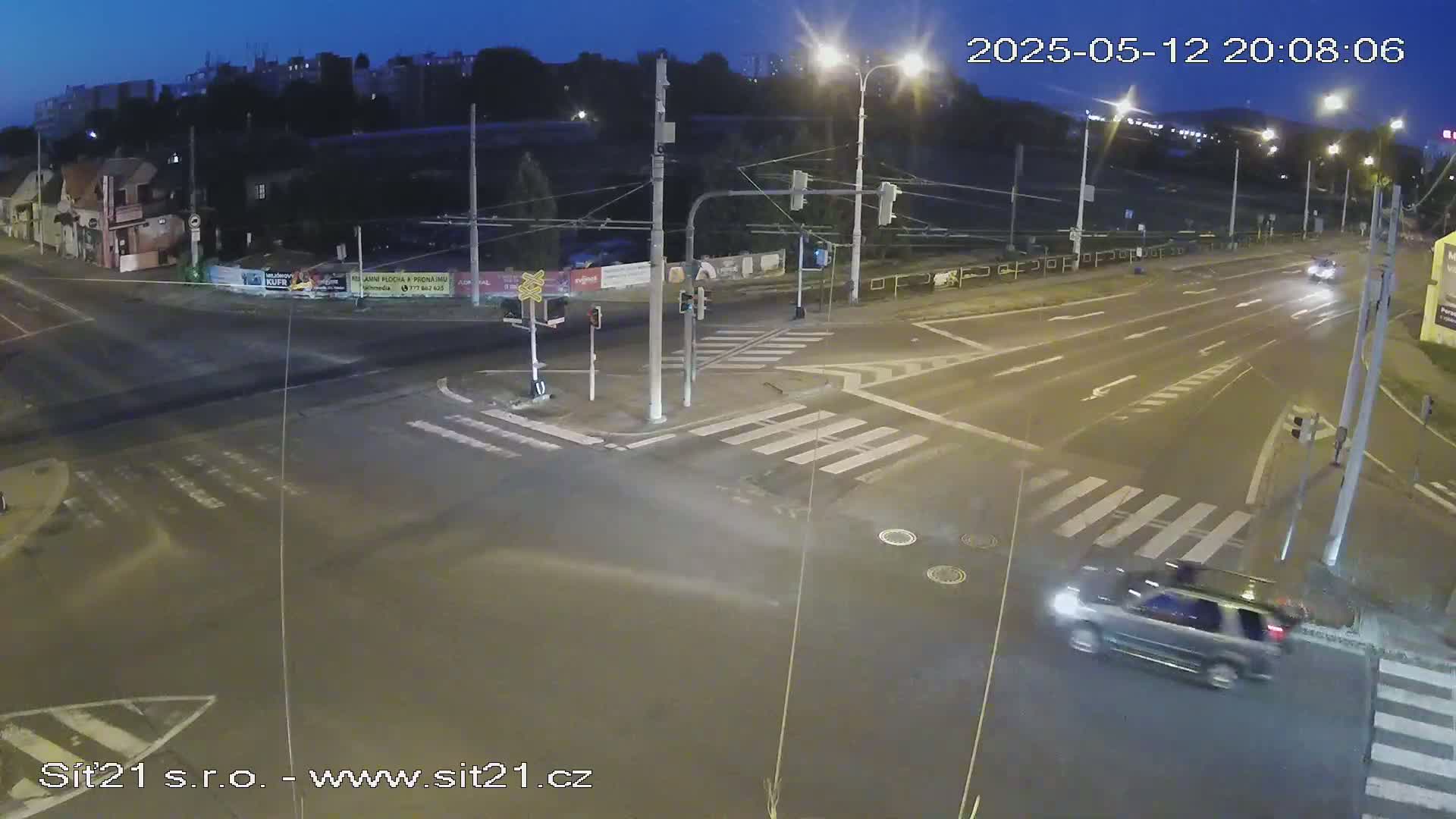 A nighttime, street-level view of a dark, empty intersection with a few cars and traffic lights under a twilight sky.