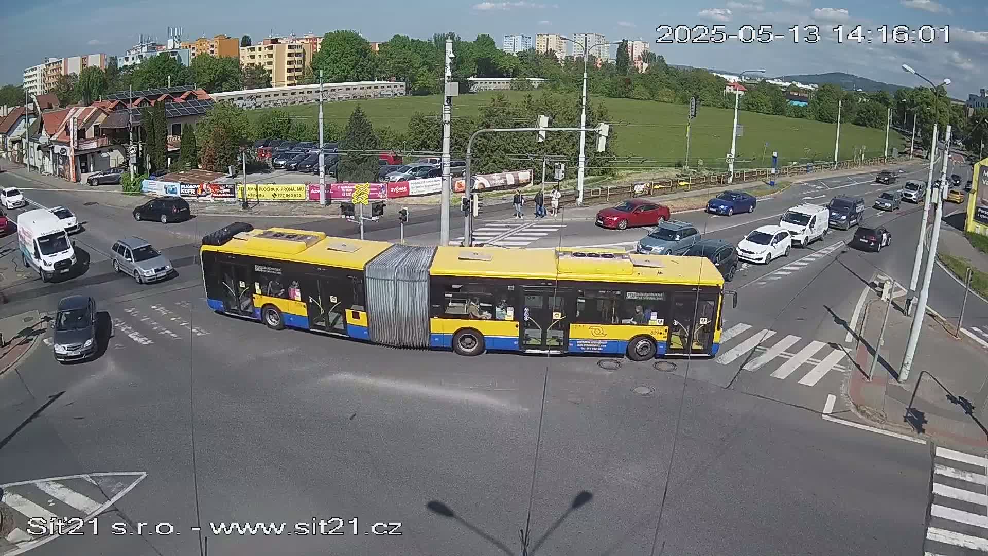 A yellow articulated bus is at a crossroads on a sunny day with several cars and pedestrians also present.