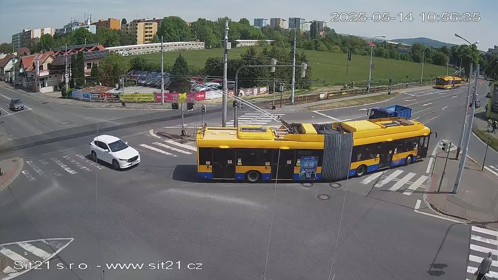 A yellow trolleybus and a white car are at a crossroads on a sunny day, with other vehicles and buildings visible in the background.