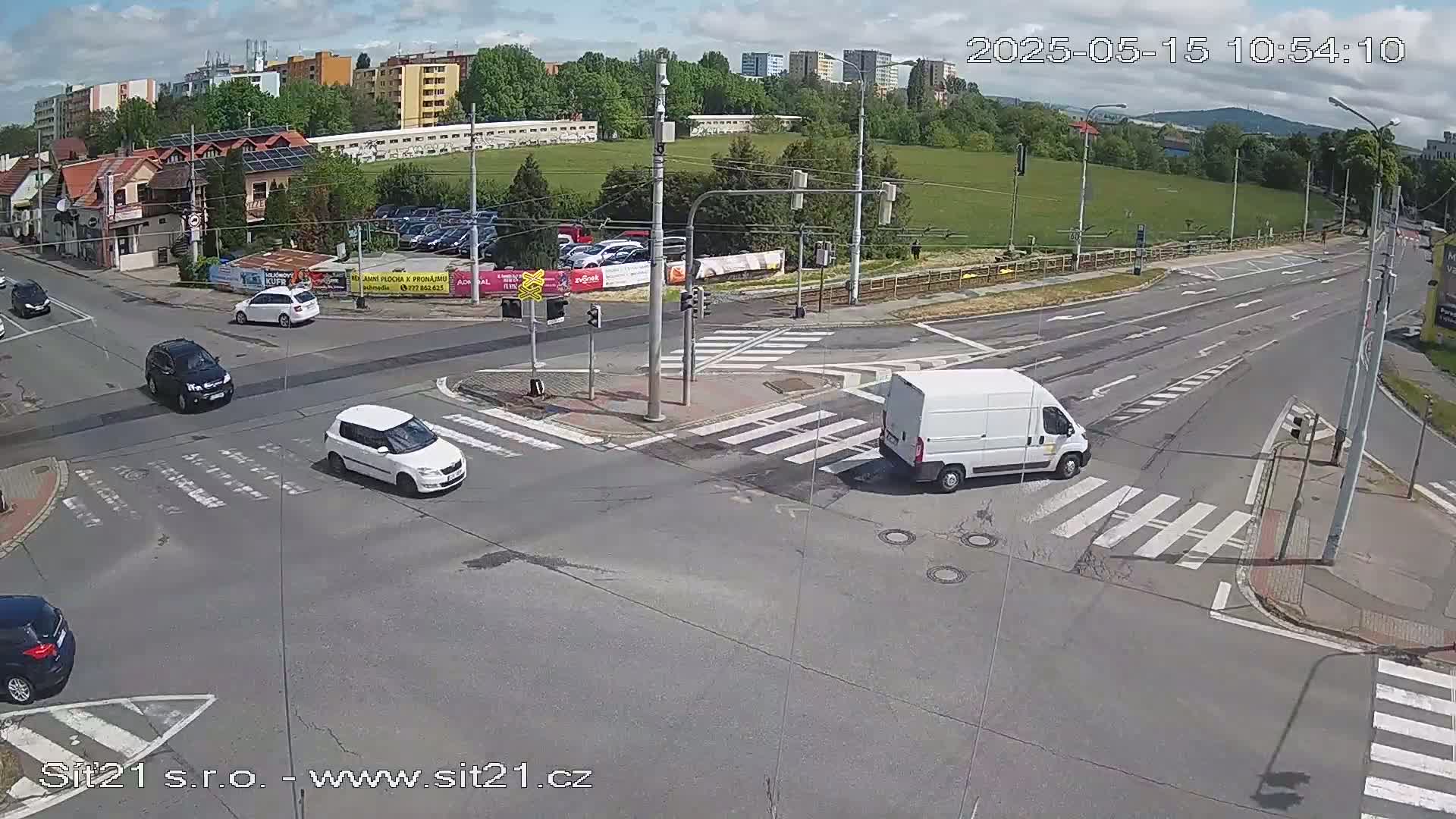 A mostly sunny day at a multi-lane road intersection with several cars and a van, where crosswalks and traffic lights are visible.