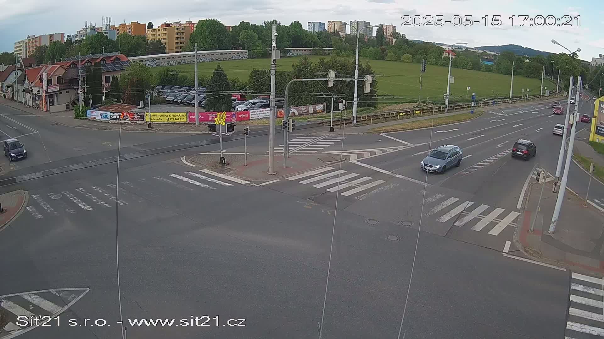 A mostly cloudy day shows a multi-lane road intersection with several cars, a pedestrian crossing, and buildings in the background.