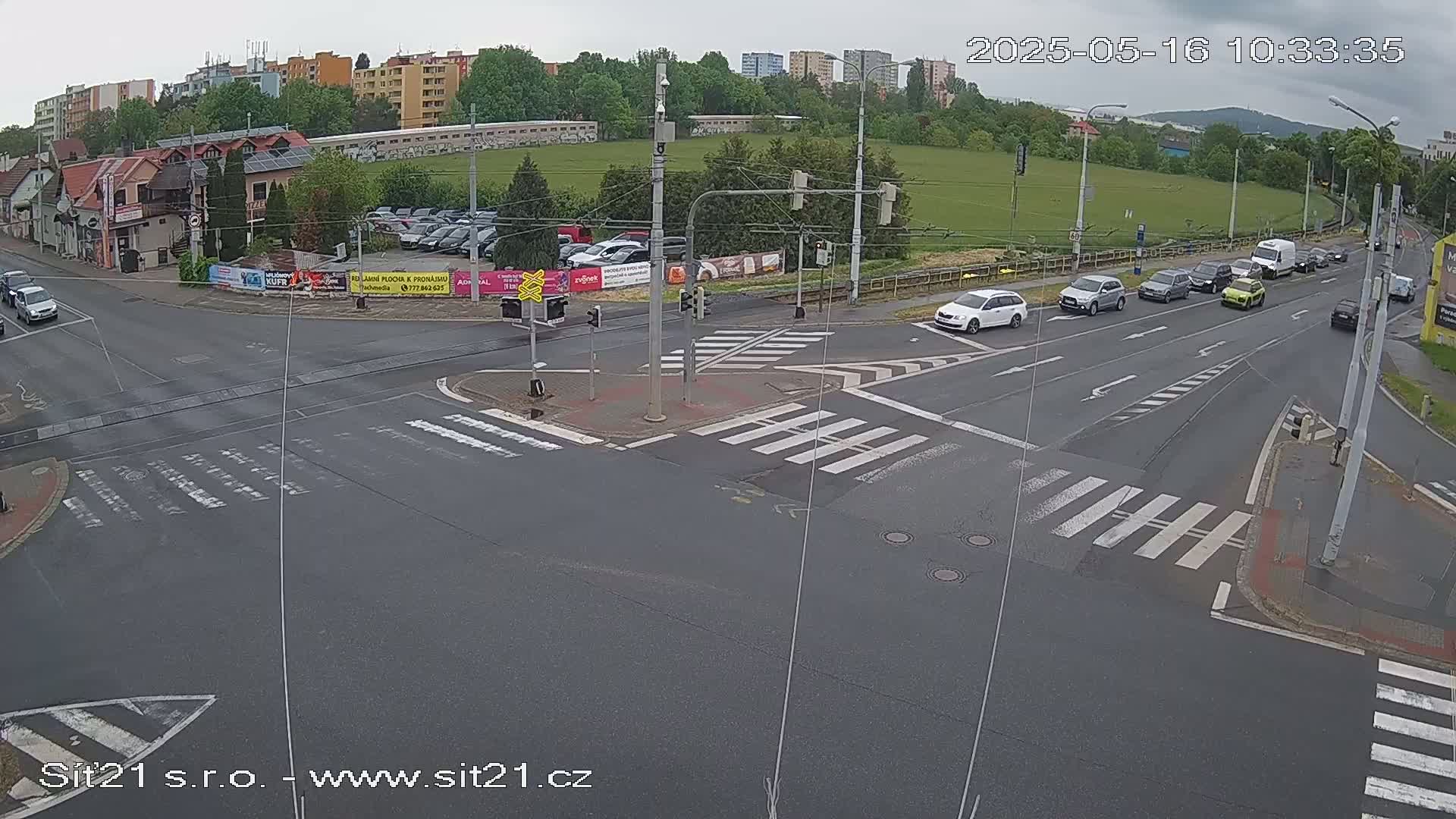 A multi-lane road intersection with several cars and a tram line, under an overcast sky.