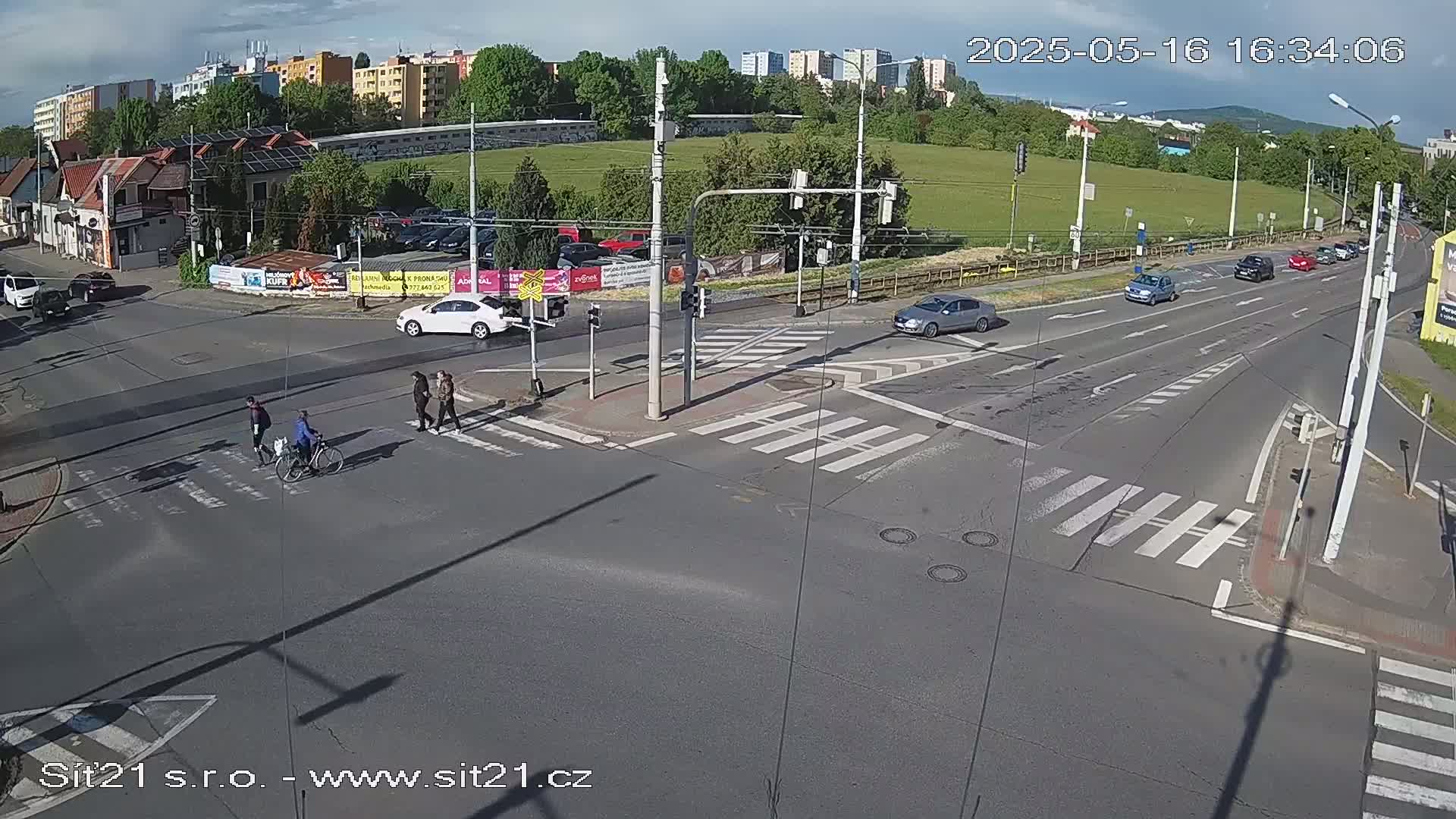 A sunny day at a multi-lane intersection shows pedestrians crossing a crosswalk and several cars driving on the road, with some parked cars and buildings in the background.