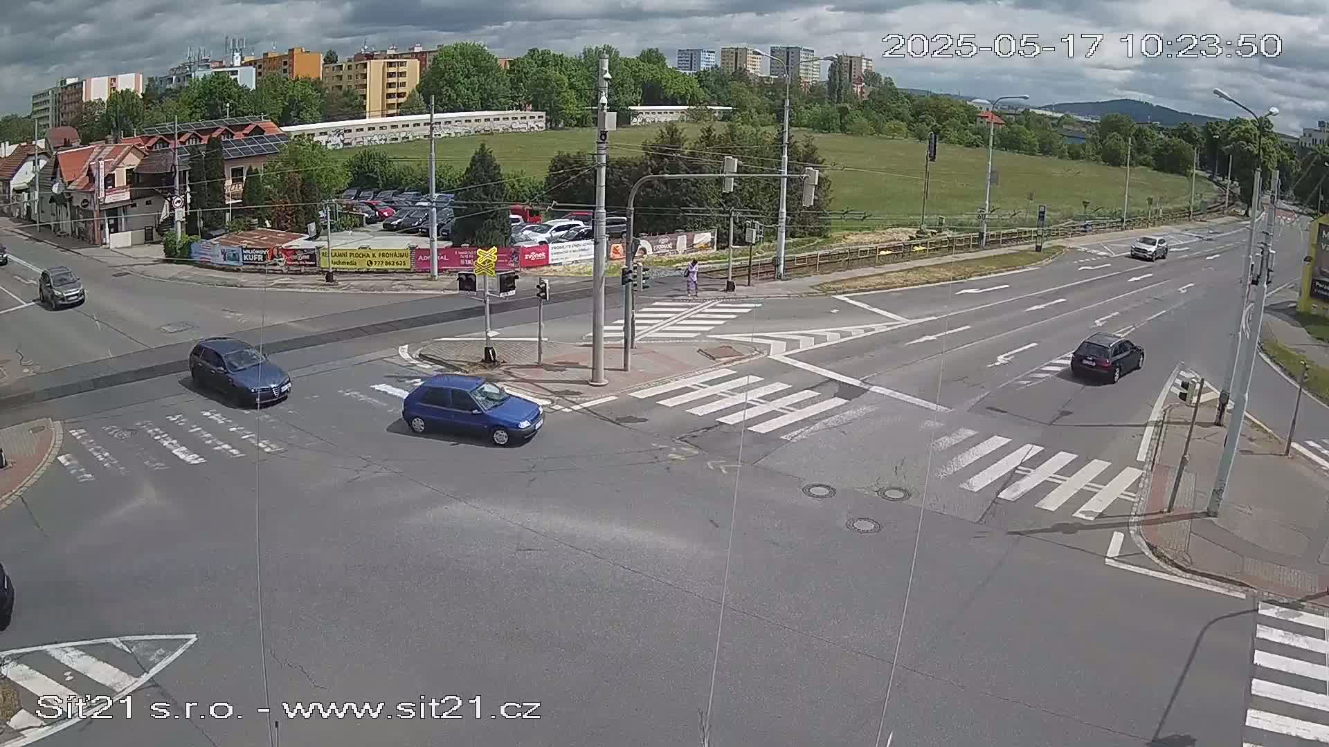 A partly cloudy day shows several cars at a multi-lane intersection with crosswalks, surrounded by buildings and green spaces.