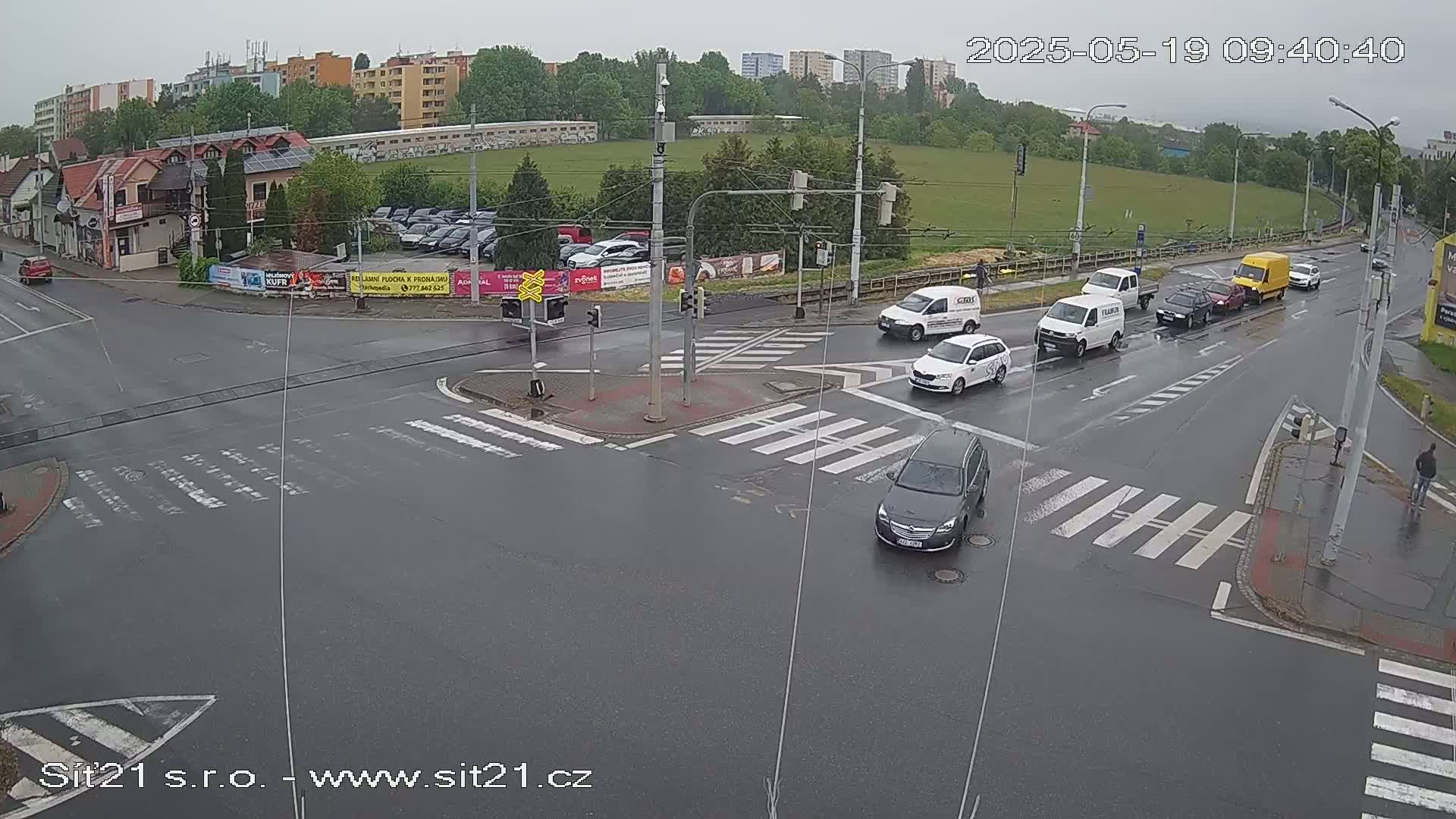 A wet, overcast day at a multi-lane intersection where several cars and vans are stopped and moving, with pedestrians crossing at marked crosswalks.