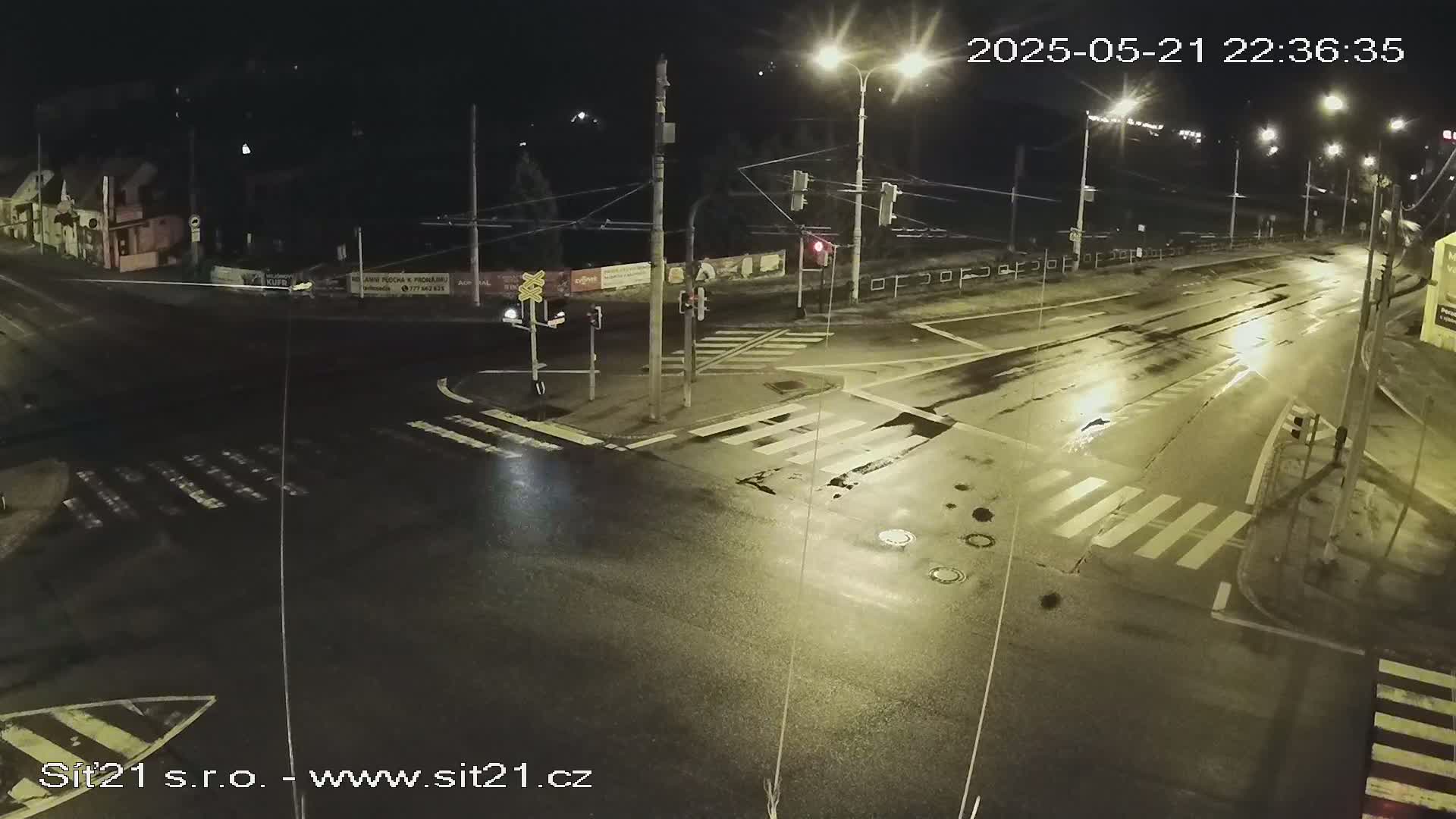 A wet, nighttime street intersection with traffic lights and crosswalks is illuminated by streetlights.