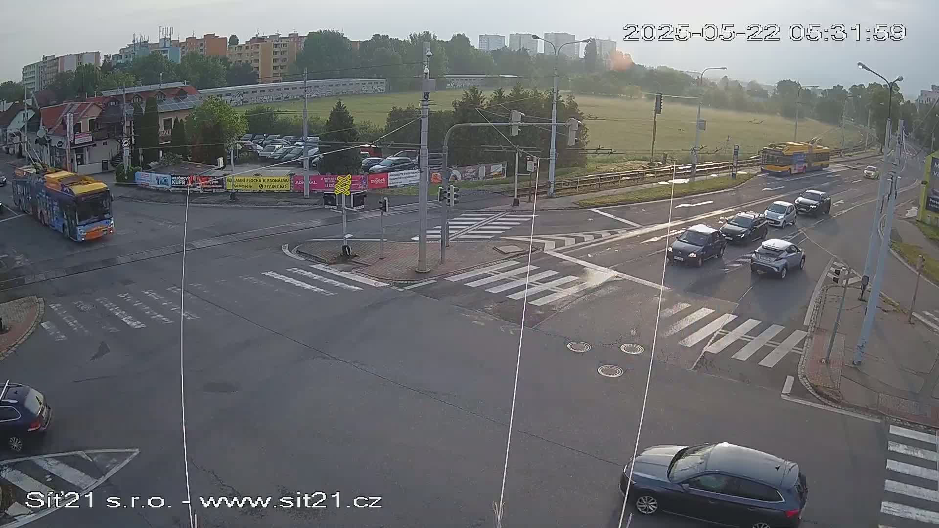 A slightly hazy morning scene shows a multi-lane intersection with several cars and two buses at various points, with crosswalks and traffic signals.