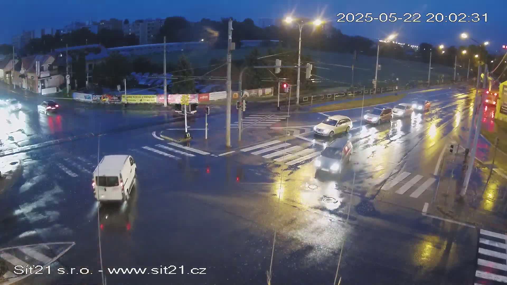 Several cars are driving through a wet intersection at night under streetlights.