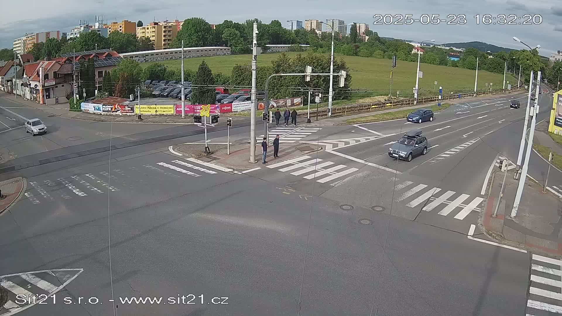 A partly cloudy day shows a street intersection with several cars and pedestrians at a crosswalk.