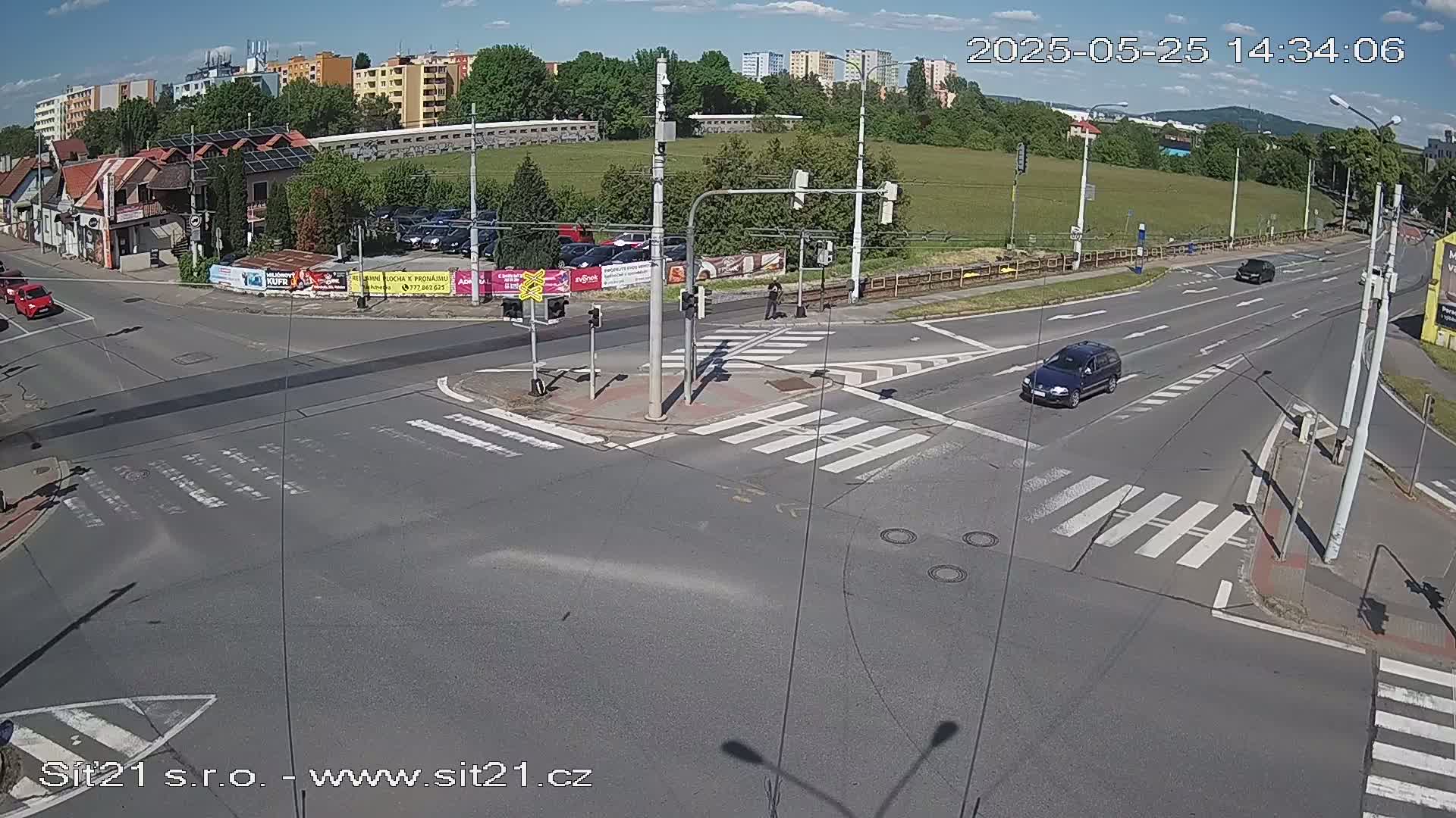 A sunny day at a multi-lane intersection with traffic lights, pedestrian crosswalks, and several cars; buildings and greenery are visible in the background.