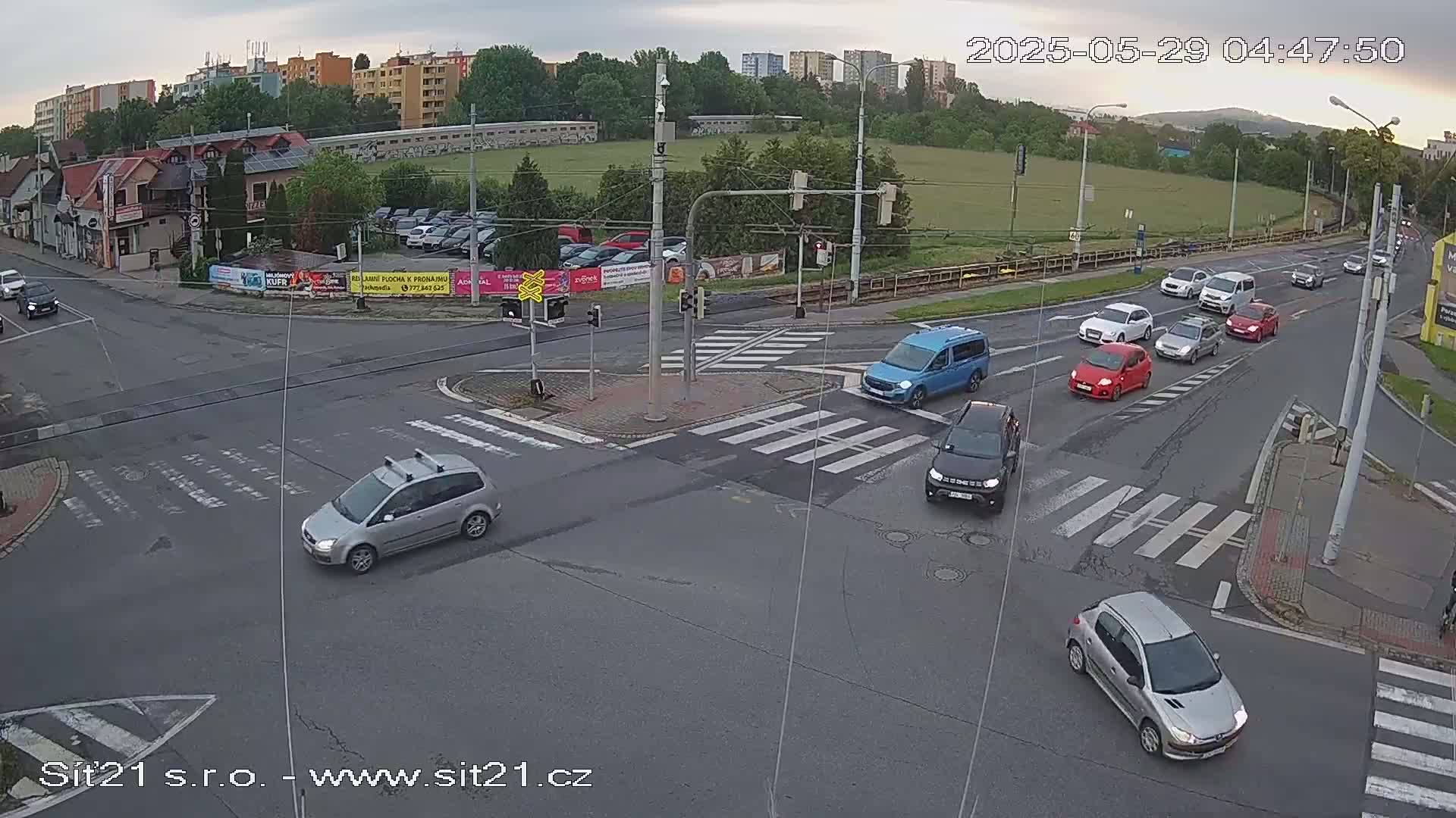 A slightly overcast outdoor scene shows several cars navigating a multi-lane intersection with crosswalks and traffic signals.