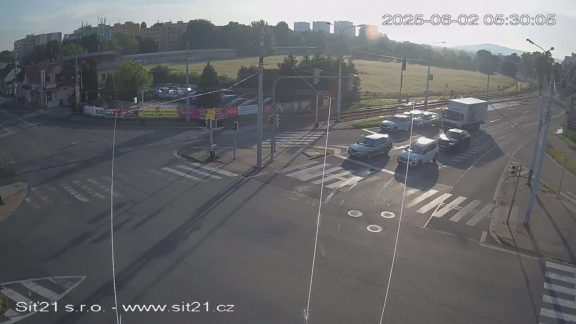 A sunny overhead view of a multi-lane road intersection with several cars and a truck navigating the crosswalks and turning lanes.