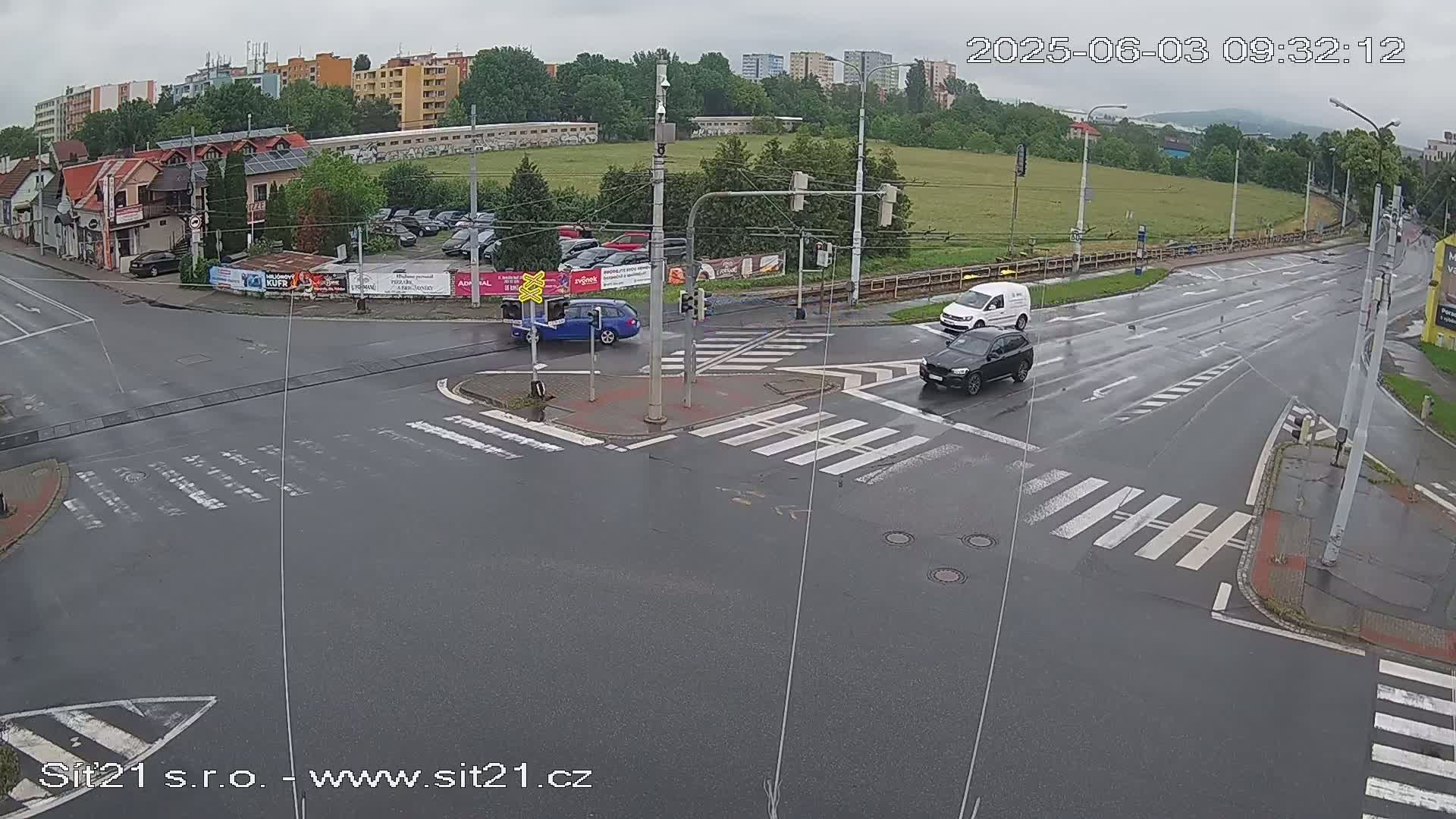 A wet, overcast road intersection shows several cars navigating a crosswalk and traffic signals under a gray sky.