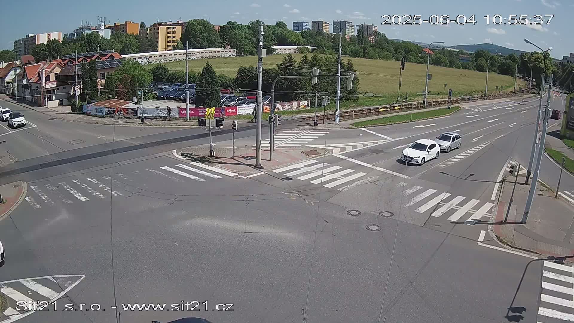 A sunny day at a multi-lane road intersection with crosswalks, showing two light-colored cars and buildings in the background.