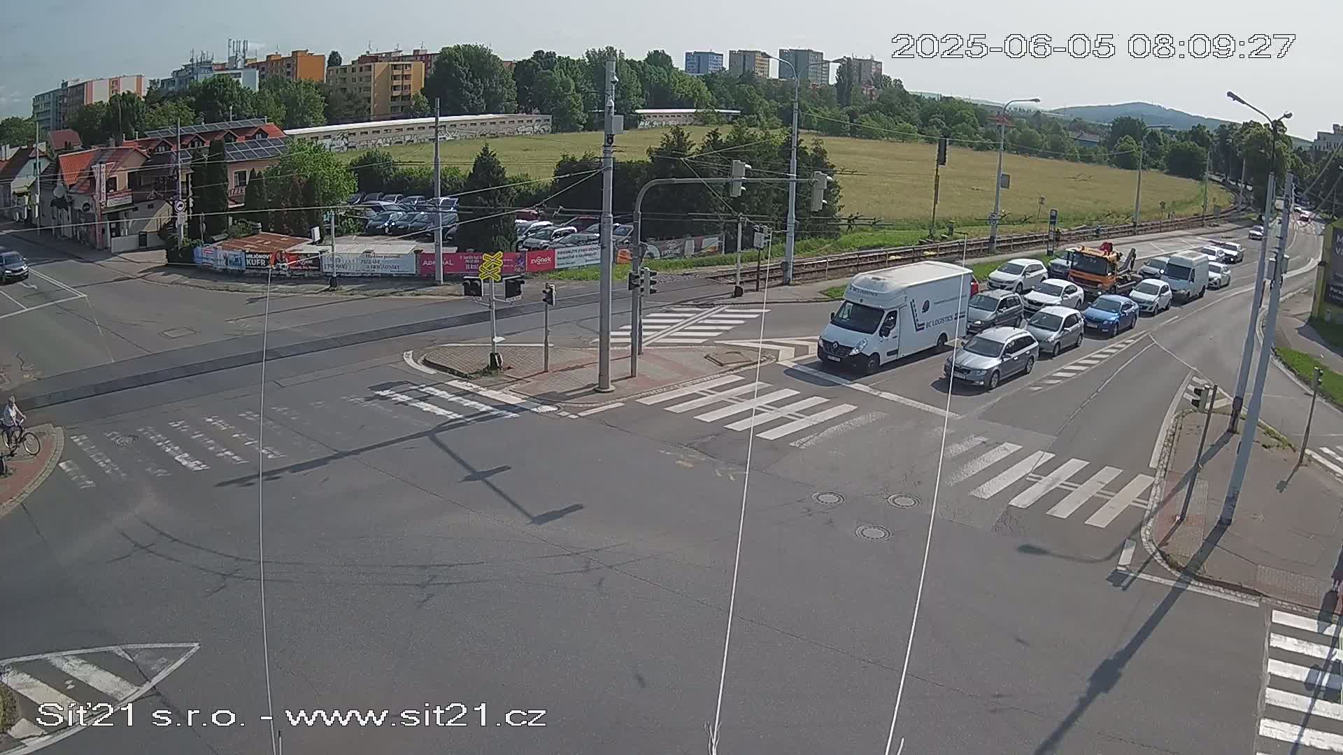 A sunny day at a multi-lane intersection shows several cars stopped at a traffic light, with a cyclist riding a bike across a crosswalk, and a white van and a tow truck visible in the scene.