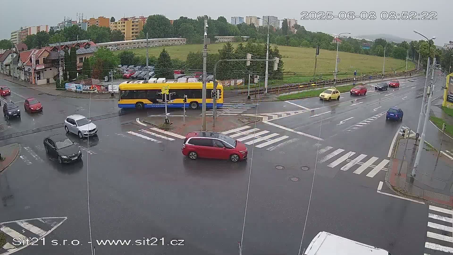 A wet street intersection shows several cars and a yellow bus, with a background of buildings and trees under an overcast sky.