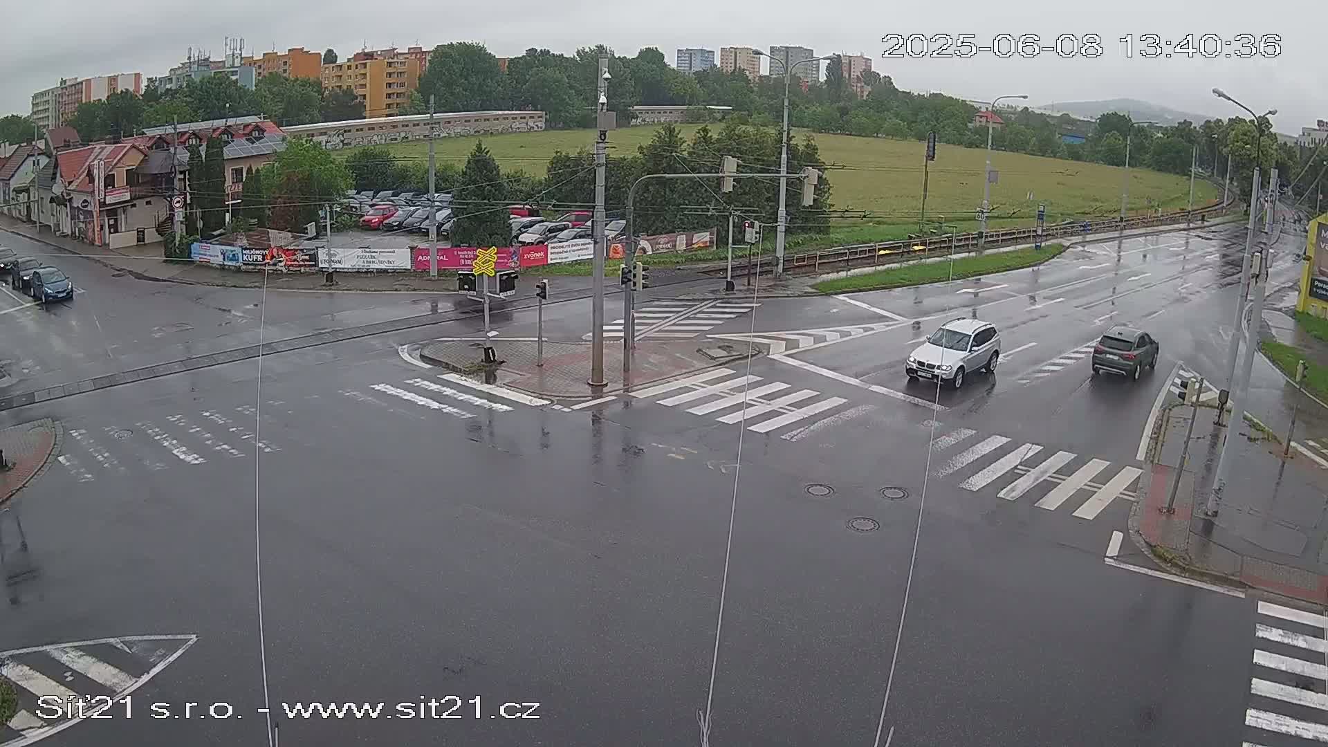 A wet, rainy intersection shows several cars navigating a crosswalk and roads under a grey sky.