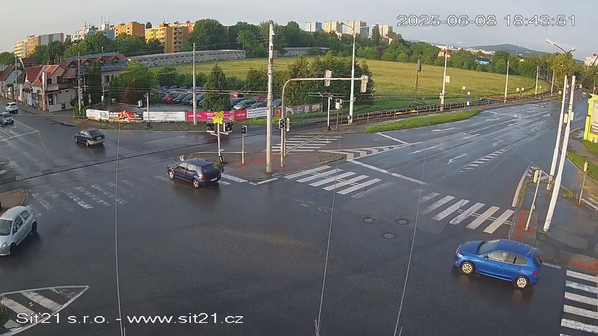 A wet road intersection with several cars, buildings, and trees in the background under an overcast sky.