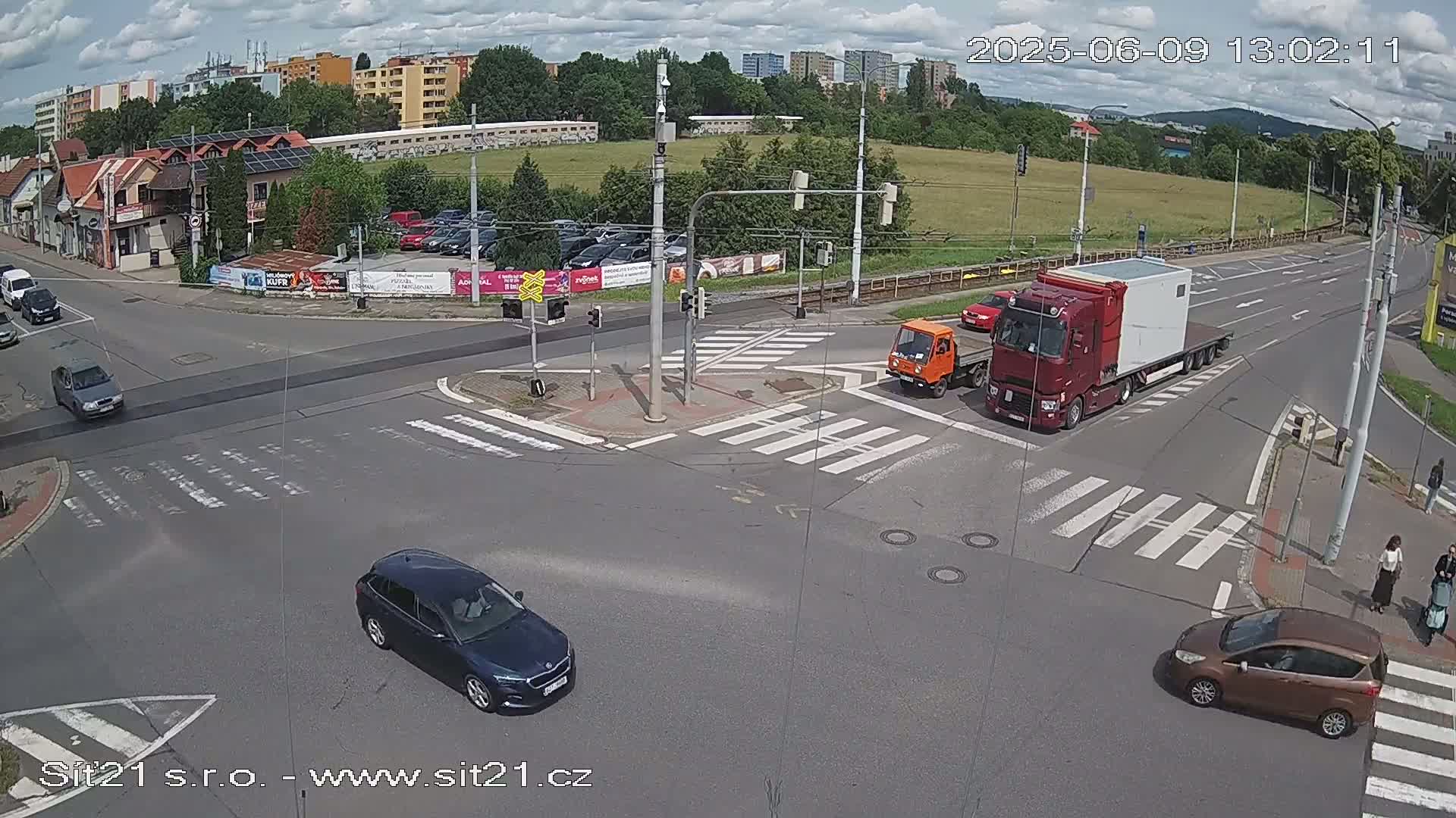 A partly cloudy day shows a multi-lane intersection with several cars and a large semi-truck navigating crosswalks and traffic signals.