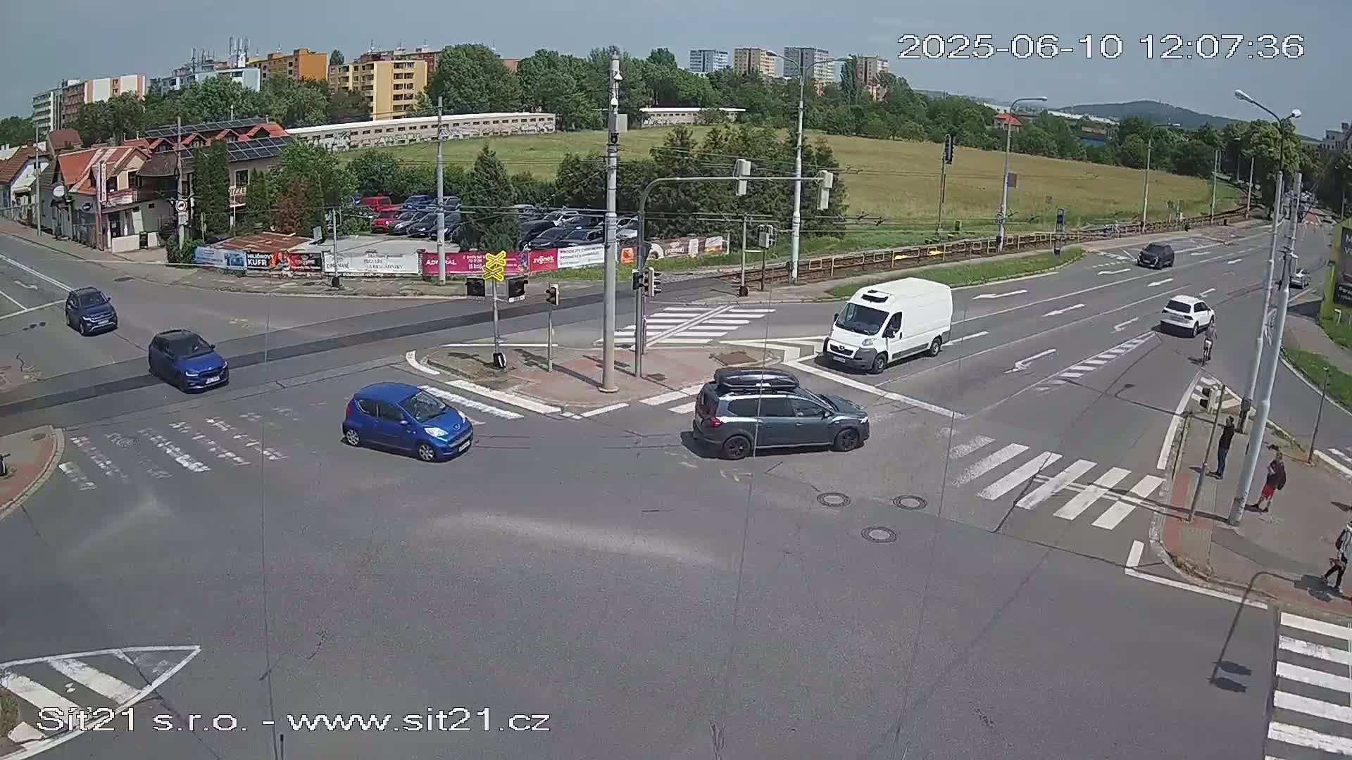 A sunny day shows a multi-lane intersection with several cars and a van, pedestrians crossing at crosswalks, and buildings and trees in the background.