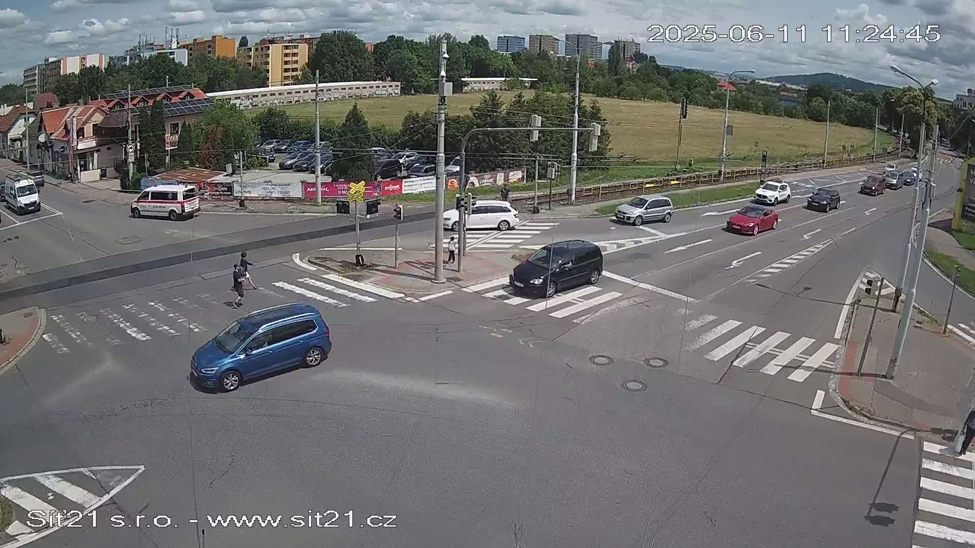 A partly sunny day shows several vehicles at a multi-lane intersection with pedestrians crossing,  nearby buildings and a grassy area visible in the background.