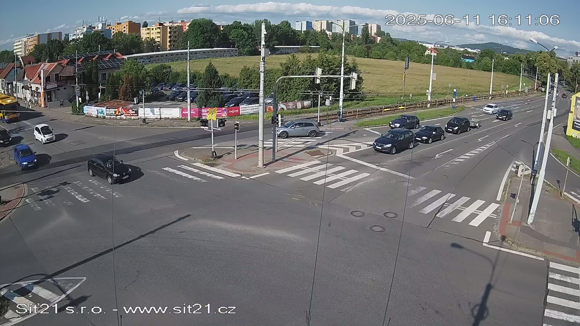 A sunny day shows several cars at a multi-lane intersection with crosswalks and traffic signals, under a partly cloudy sky.