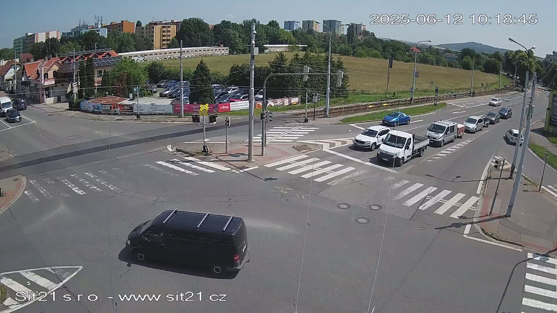 A sunny day shows a multi-lane intersection with several vehicles, including vans and cars, navigating a crosswalk and traffic signals.