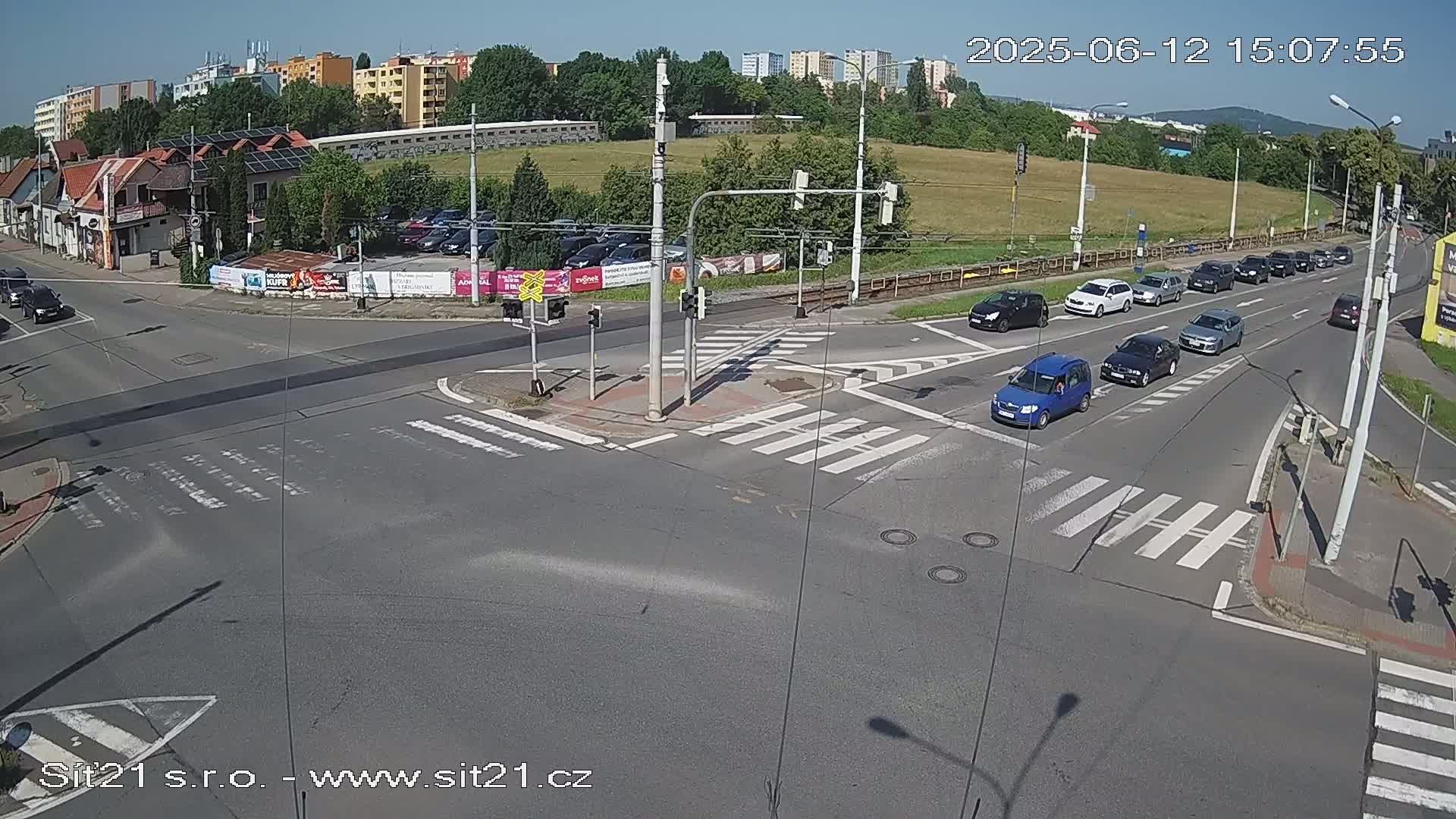 A sunny day shows a multi-lane intersection with several cars stopped at a traffic light and a few cars moving through the intersection.