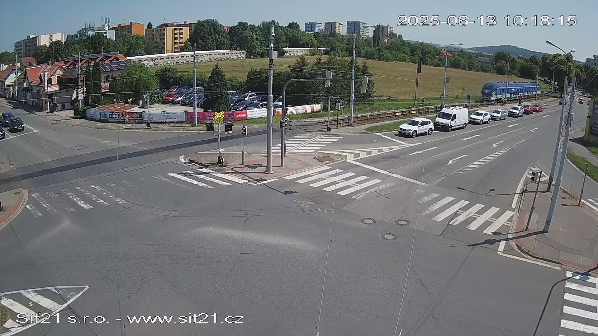 A sunny day shows a multi-lane intersection with several cars and a train passing by, near buildings and a parking lot.
