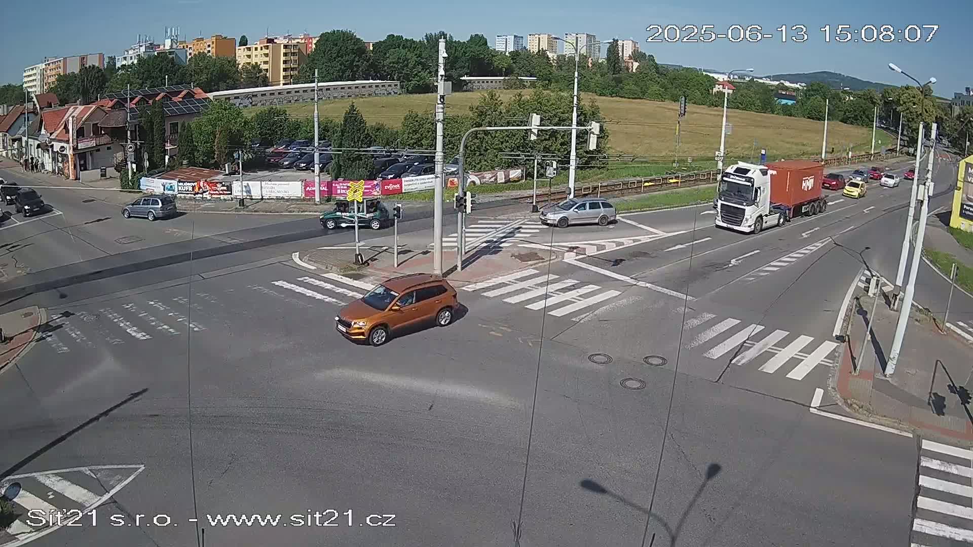 A busy intersection on a sunny day shows several cars and a large truck moving through crosswalks and traffic signals.