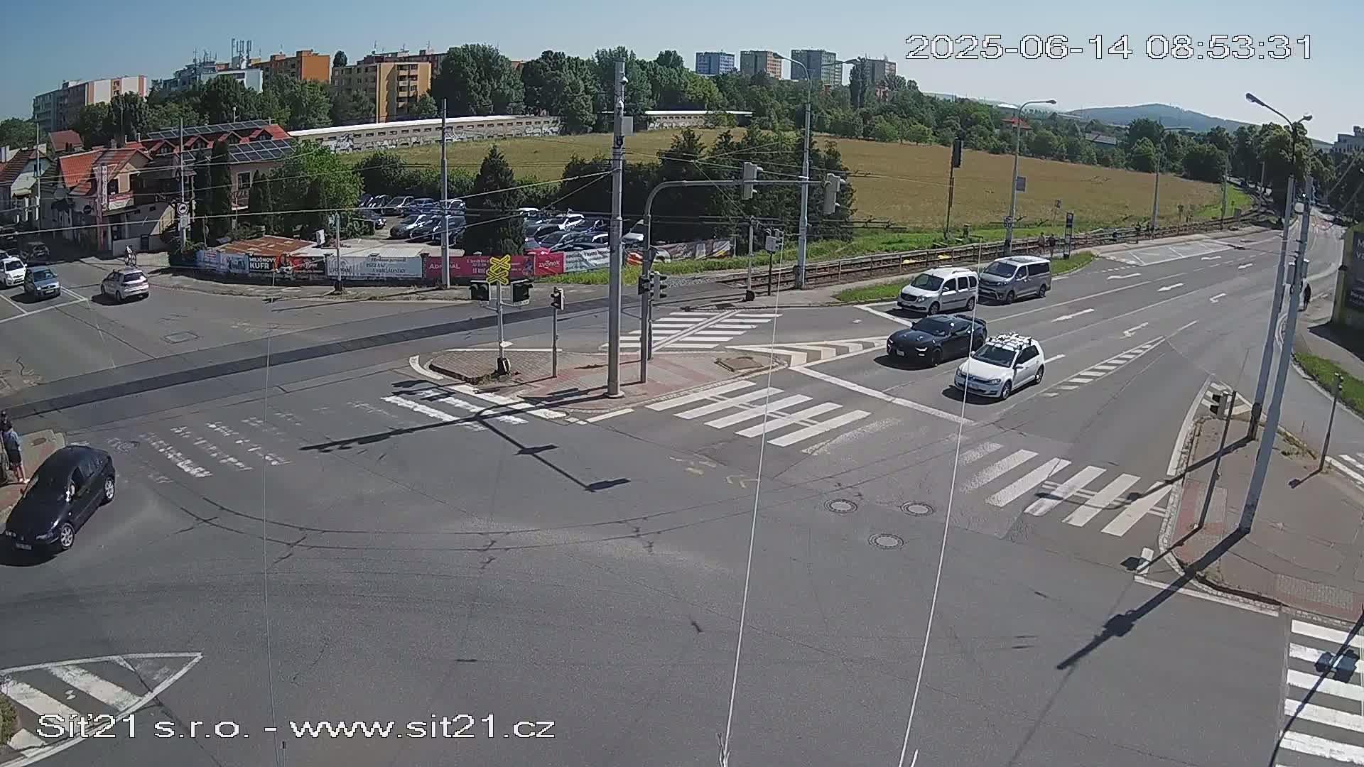 A sunny day shows a multi-lane road intersection with several cars, including a dark sedan and a white station wagon, at a crosswalk.