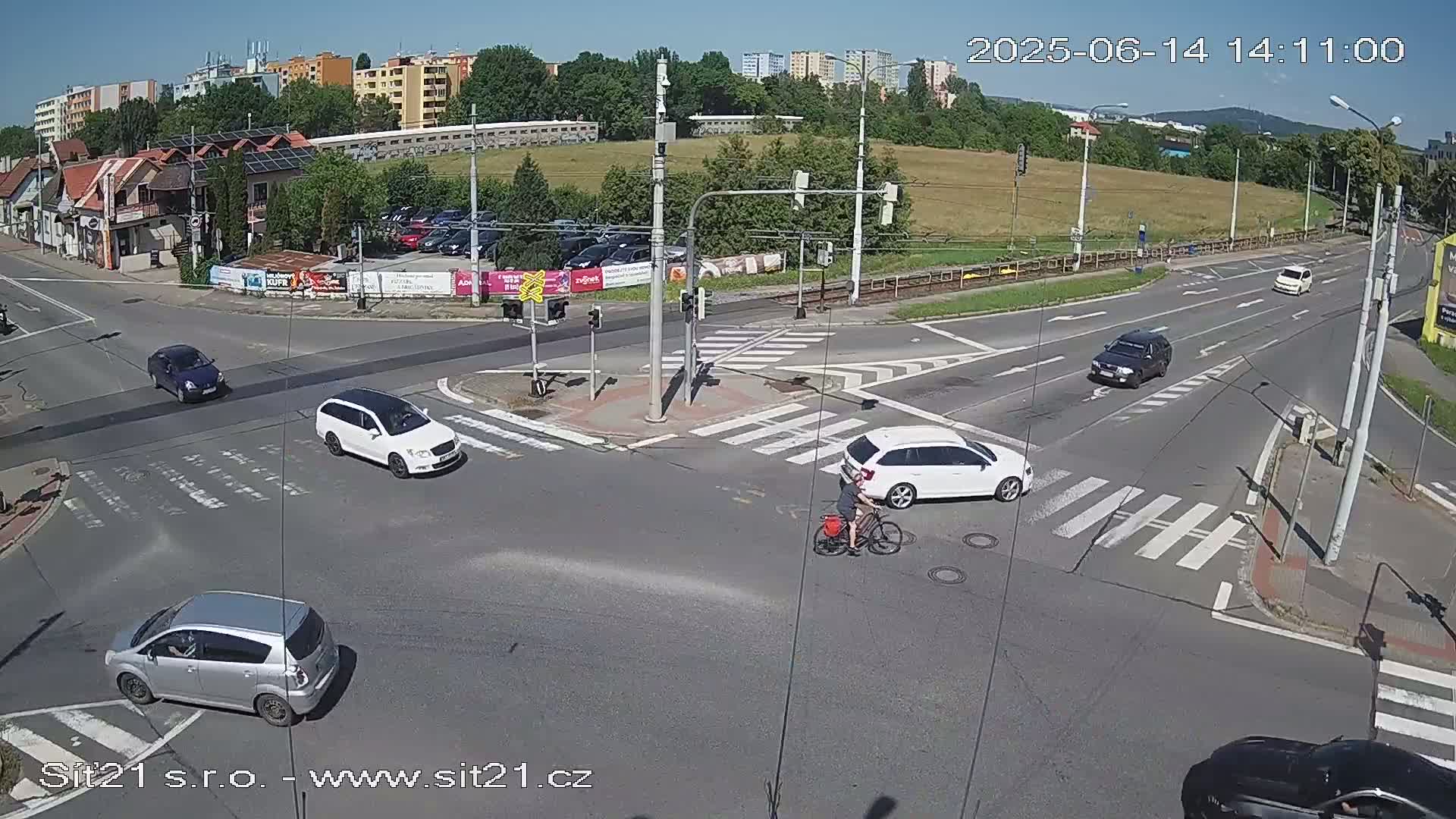 A sunny day at a multi-lane intersection shows several cars, a cyclist, and buildings in the background.