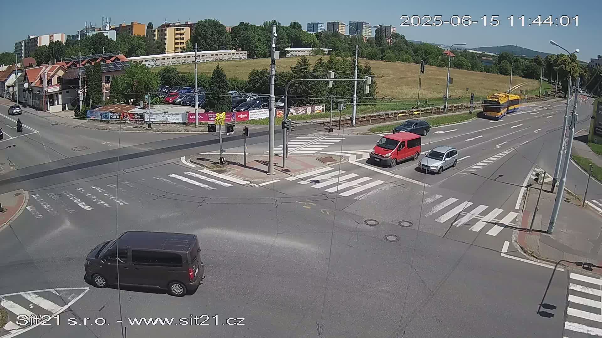 A sunny day shows a multi-lane road intersection with several vehicles, including a van, a car, and a trolleybus, near buildings and a grassy area.