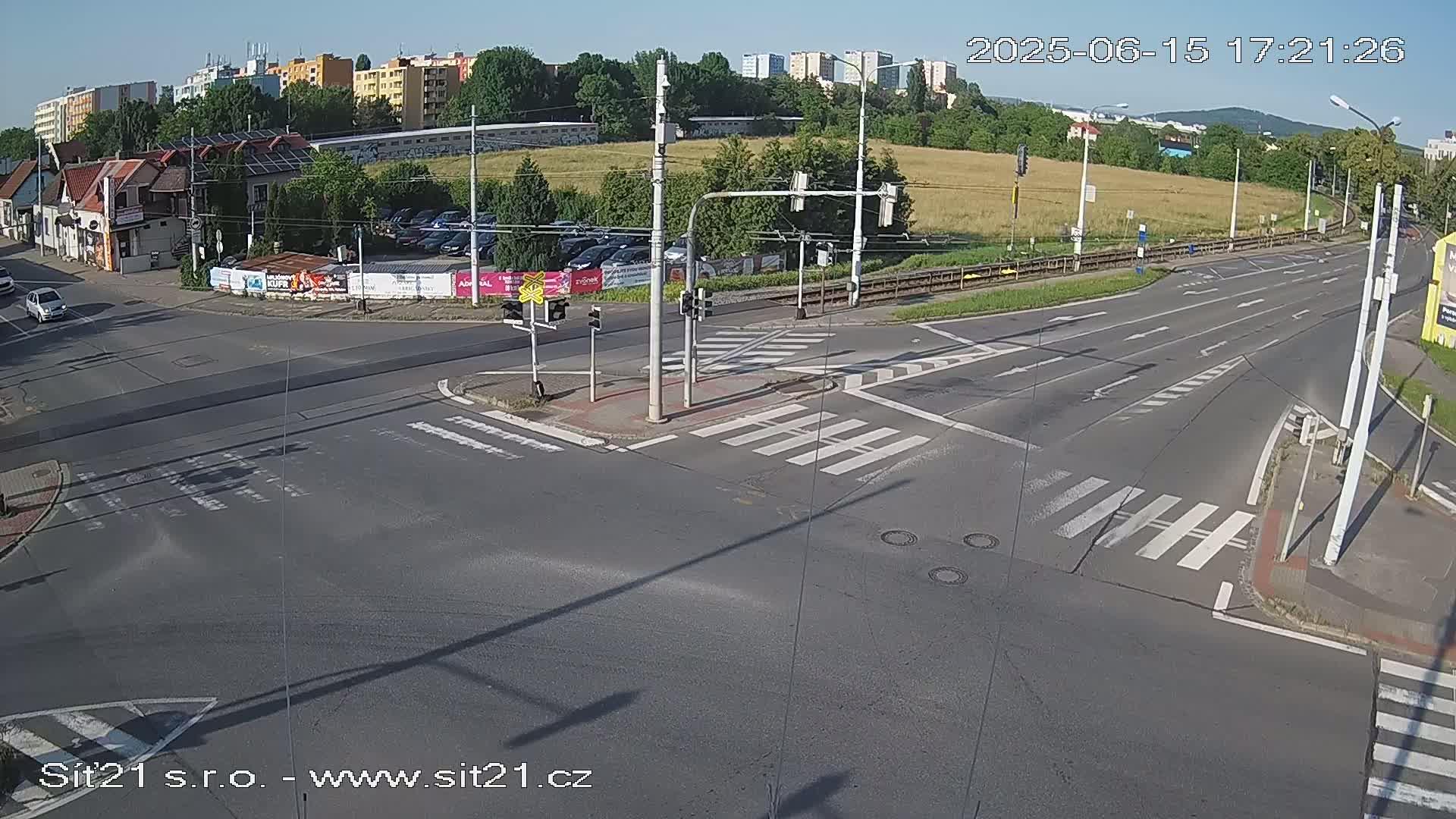 A sunny overhead view of a multi-lane road intersection with crosswalks, traffic signals, and several cars visible.
