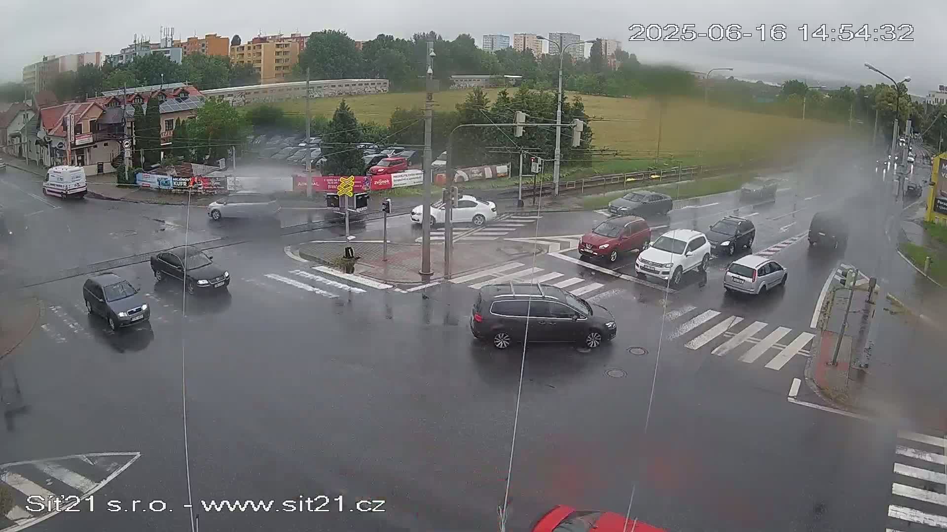 A rain-soaked intersection shows several cars navigating a crosswalk, with additional vehicles visible in the background under a cloudy sky.