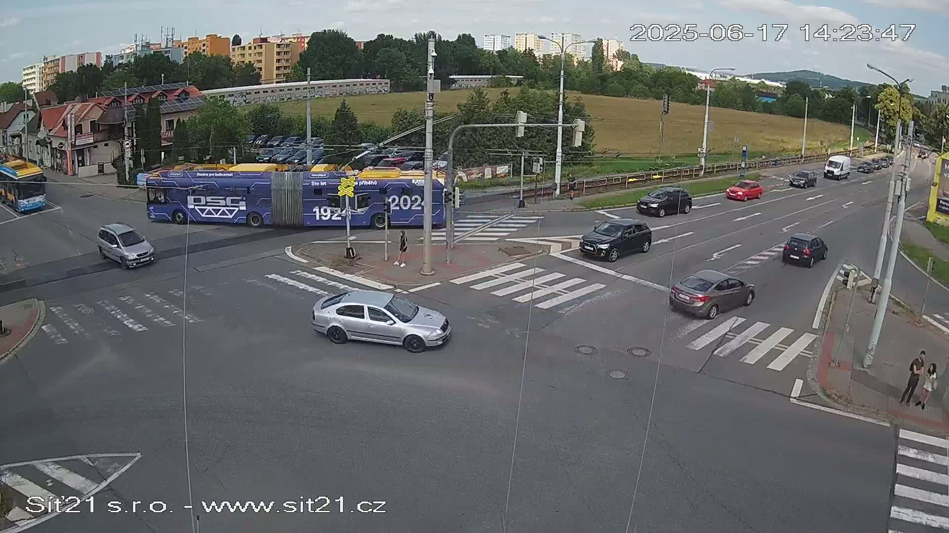 A partly sunny day shows several cars and a long, purple bus at a crossroads with pedestrian crossings.